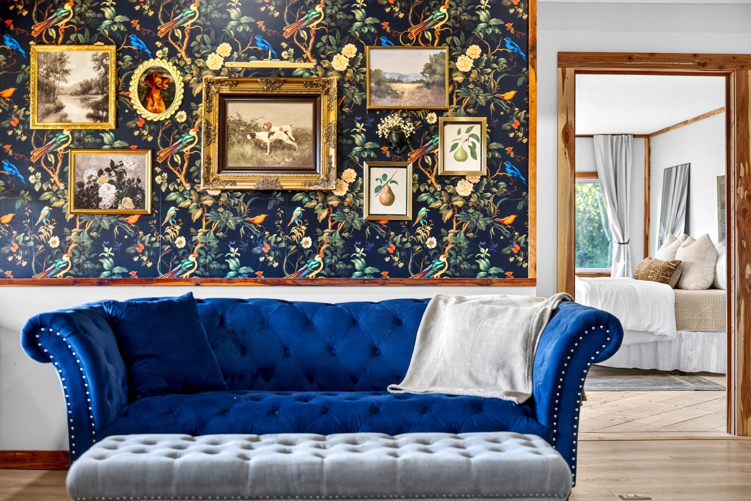 Cozy sitting area at Refuge Ranch with a navy velvet sofa, floral accent wall, curated gallery of framed artwork, and a view into the adjoining bedroom. 