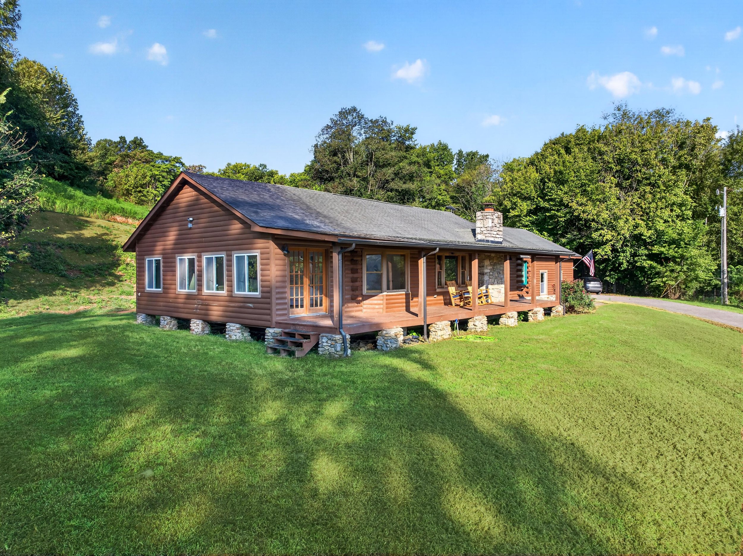 A house with brown siding and a gray roof, surrounded by green grass and trees, with a blue sky overhead.