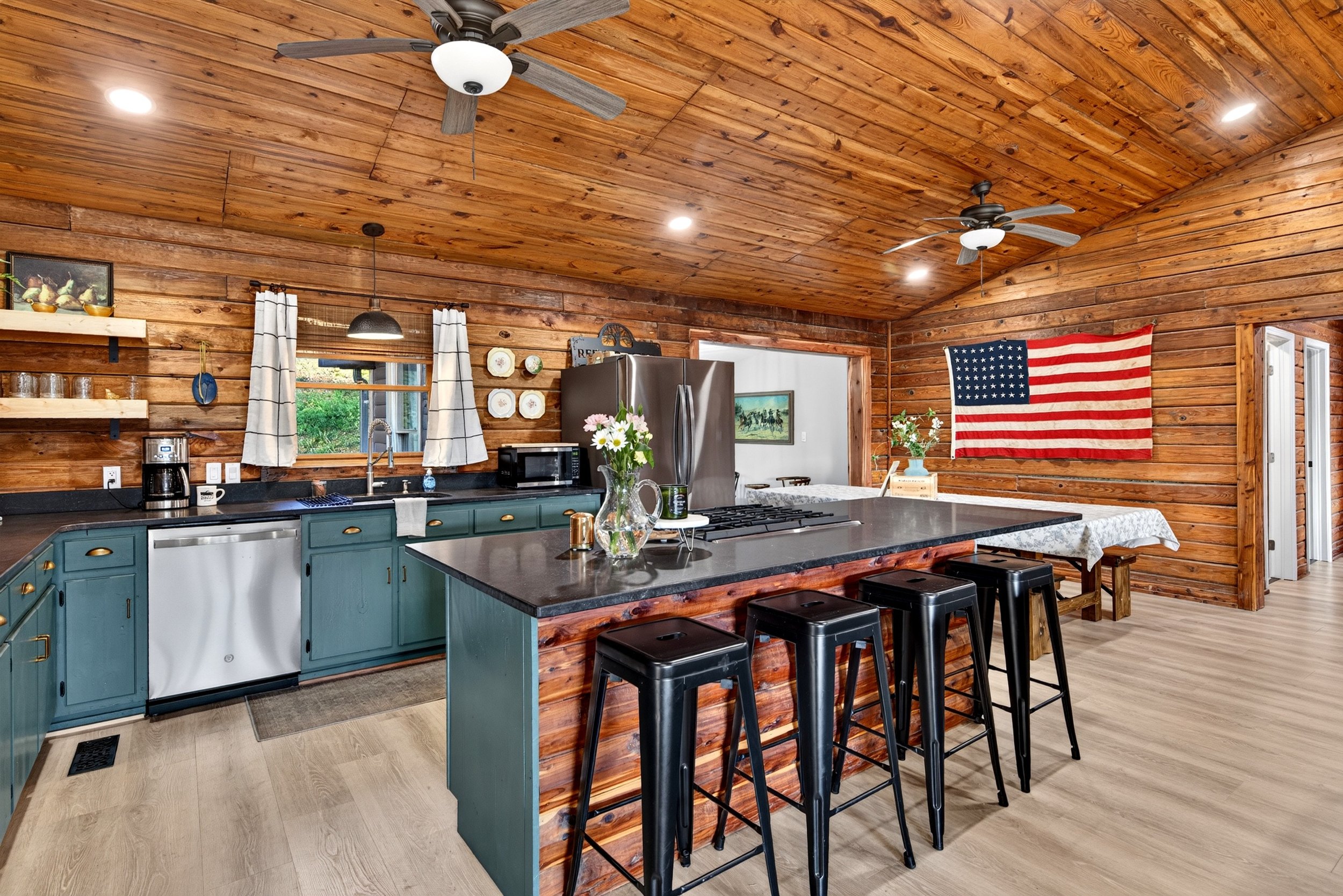 Kitchen at Refuge Ranch with warm wood-paneled walls and ceiling, green cabinetry, large island with seating, stainless steel appliances, and dinning area. 