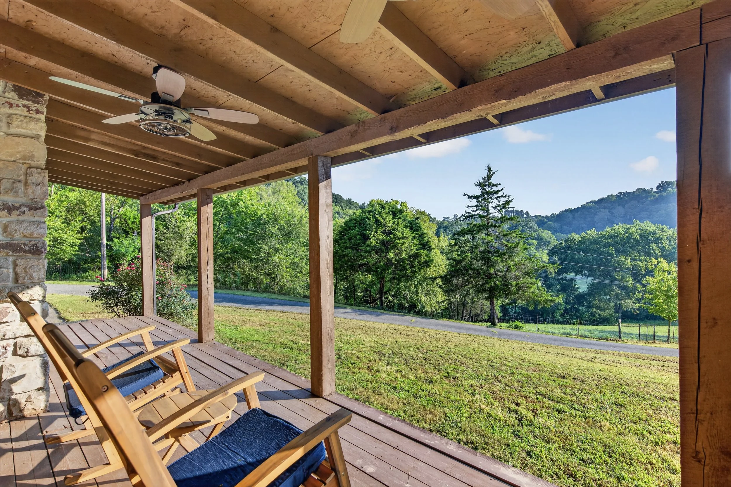 Wooden porch overlooking a grassy yard with trees and hills in the background, featuring a ceiling fan and outdoor chairs.