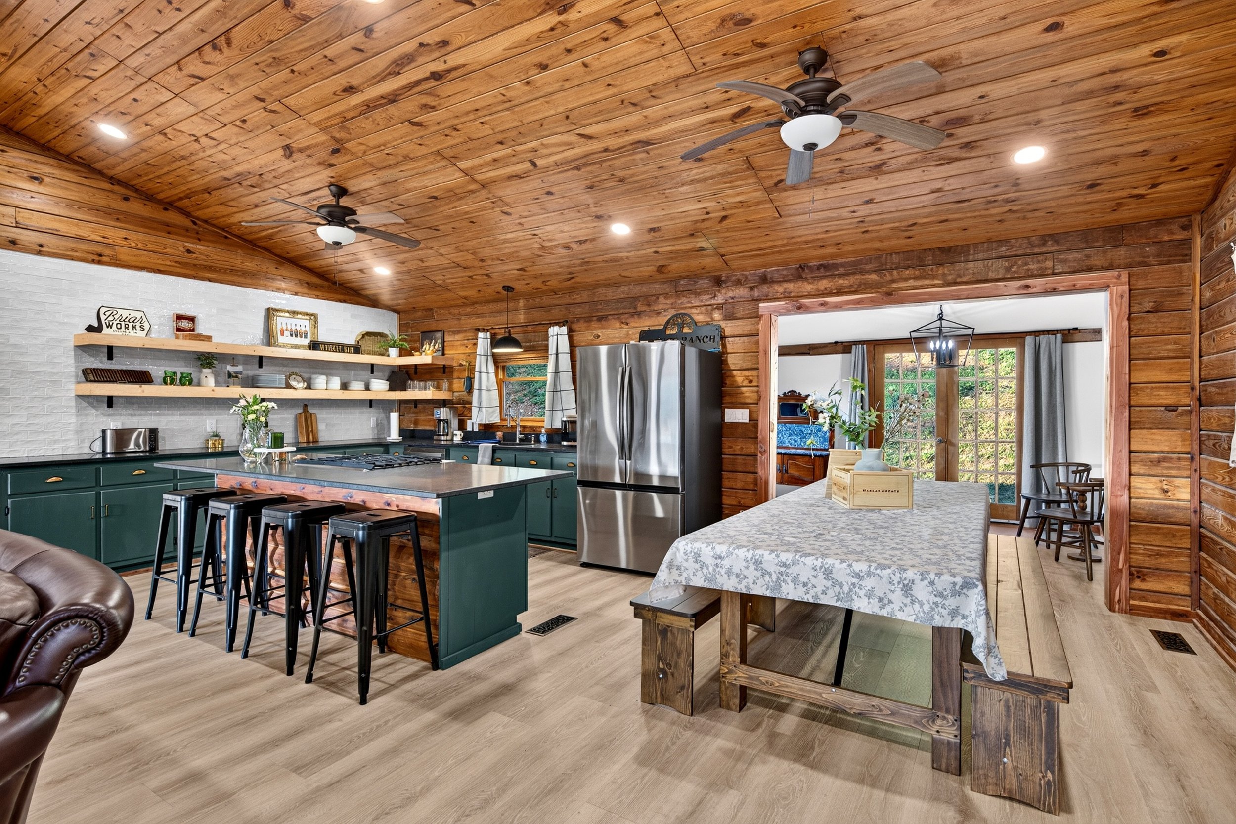 Kitchen with wooden ceiling and walls, green kitchen island with black stools, stainless steel refrigerator, open shelves with dishes and decor, window with curtains, and a dining table with a floral tablecloth.