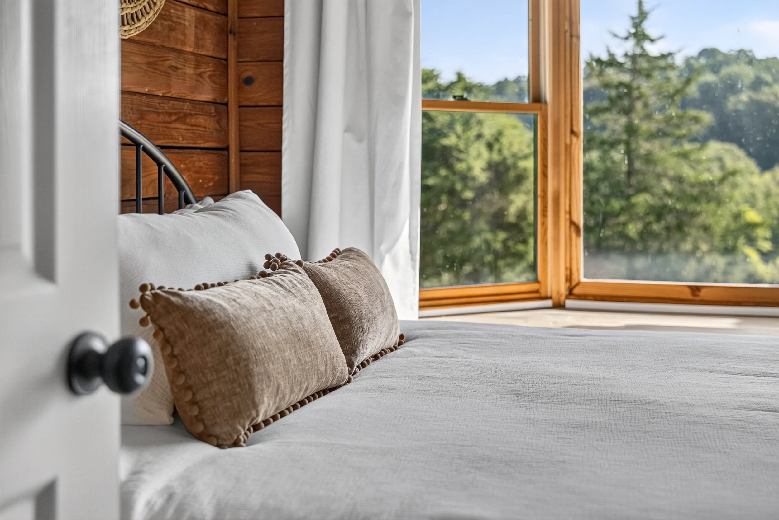 Cozy front guest bedroom at Refuge Ranch with warm wood-paneled walls and large window overlooking the trees and sunrise views.