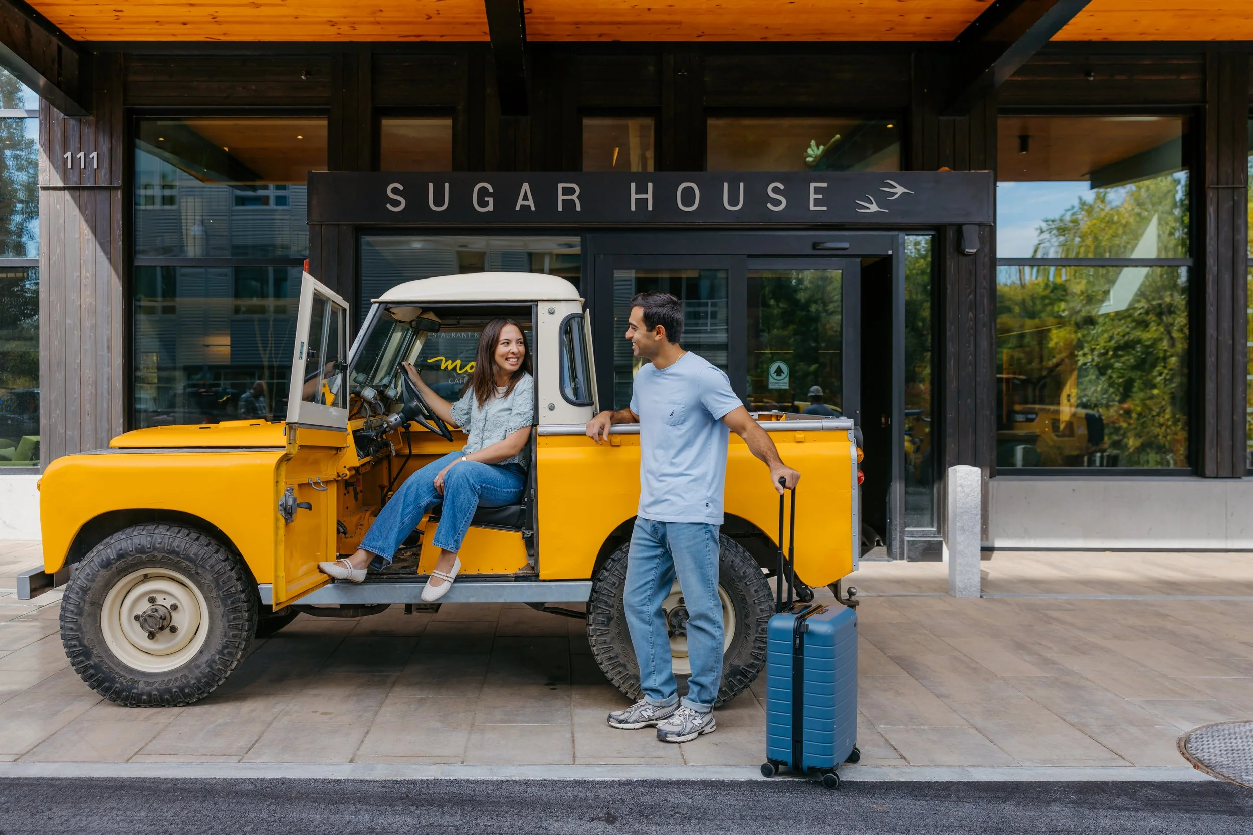 A woman sitting inside a yellow vintage jeep, smiling at a man standing beside it with luggage, in front of a building labeled 'Sugar House'.