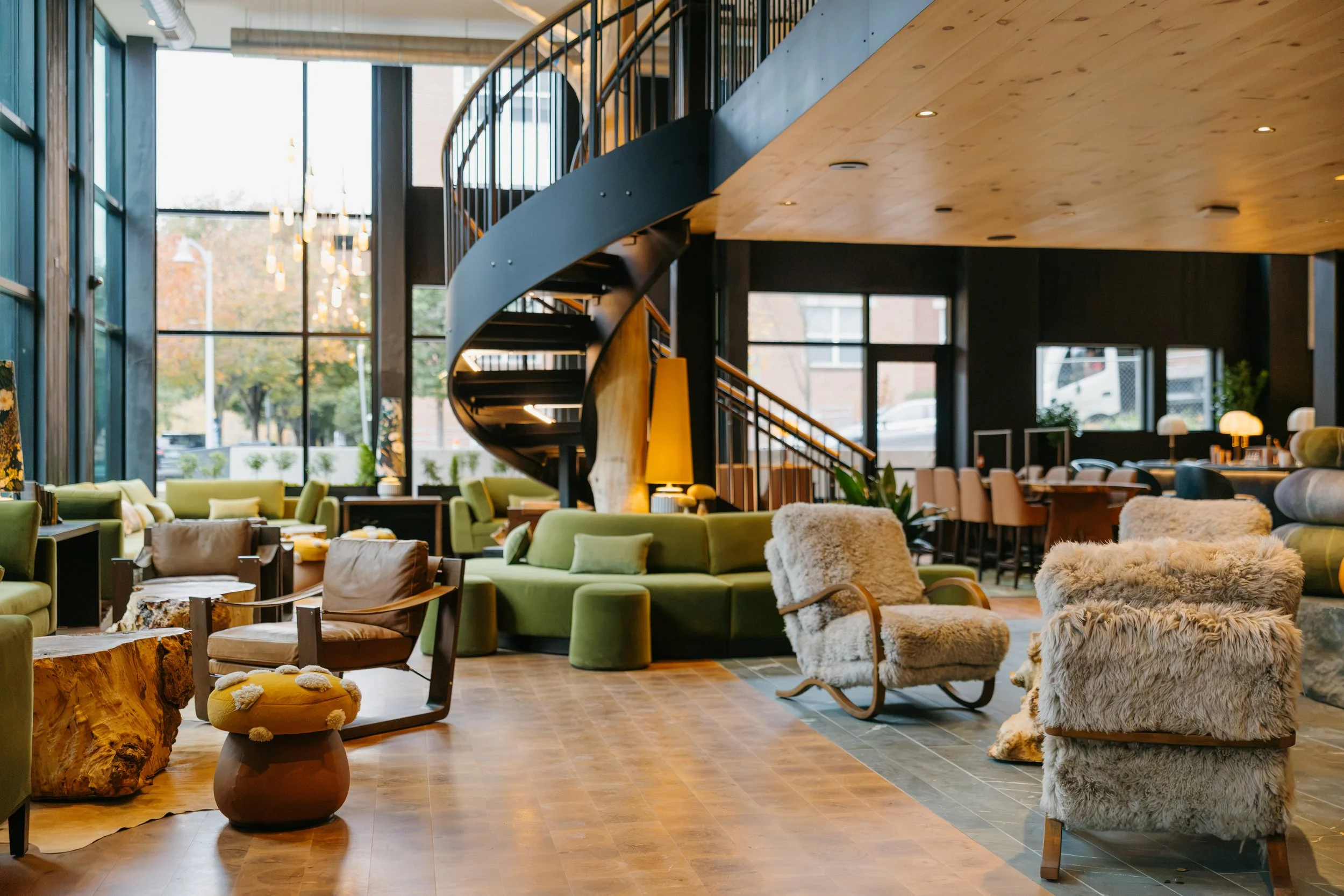 Interior of a modern lounge or lobby with large windows, a curved staircase, and various seating options including plush chairs, sofas, and armchairs.