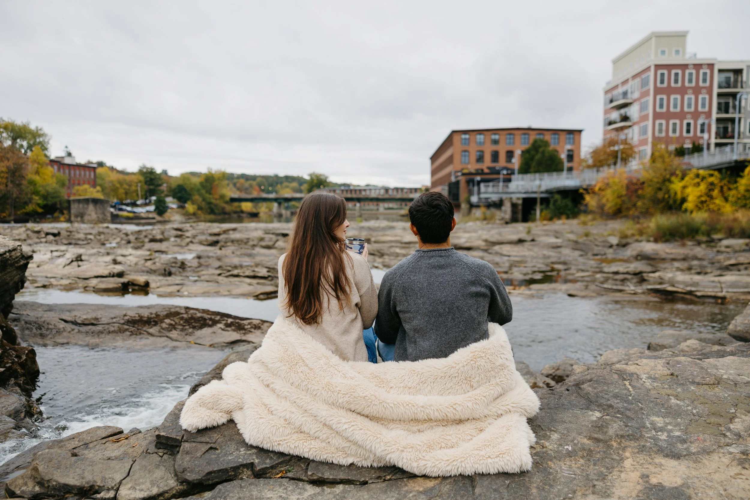 A man and woman sitting on rocks by a river, wrapped in a beige blanket, with city buildings and trees in the background on an overcast day.