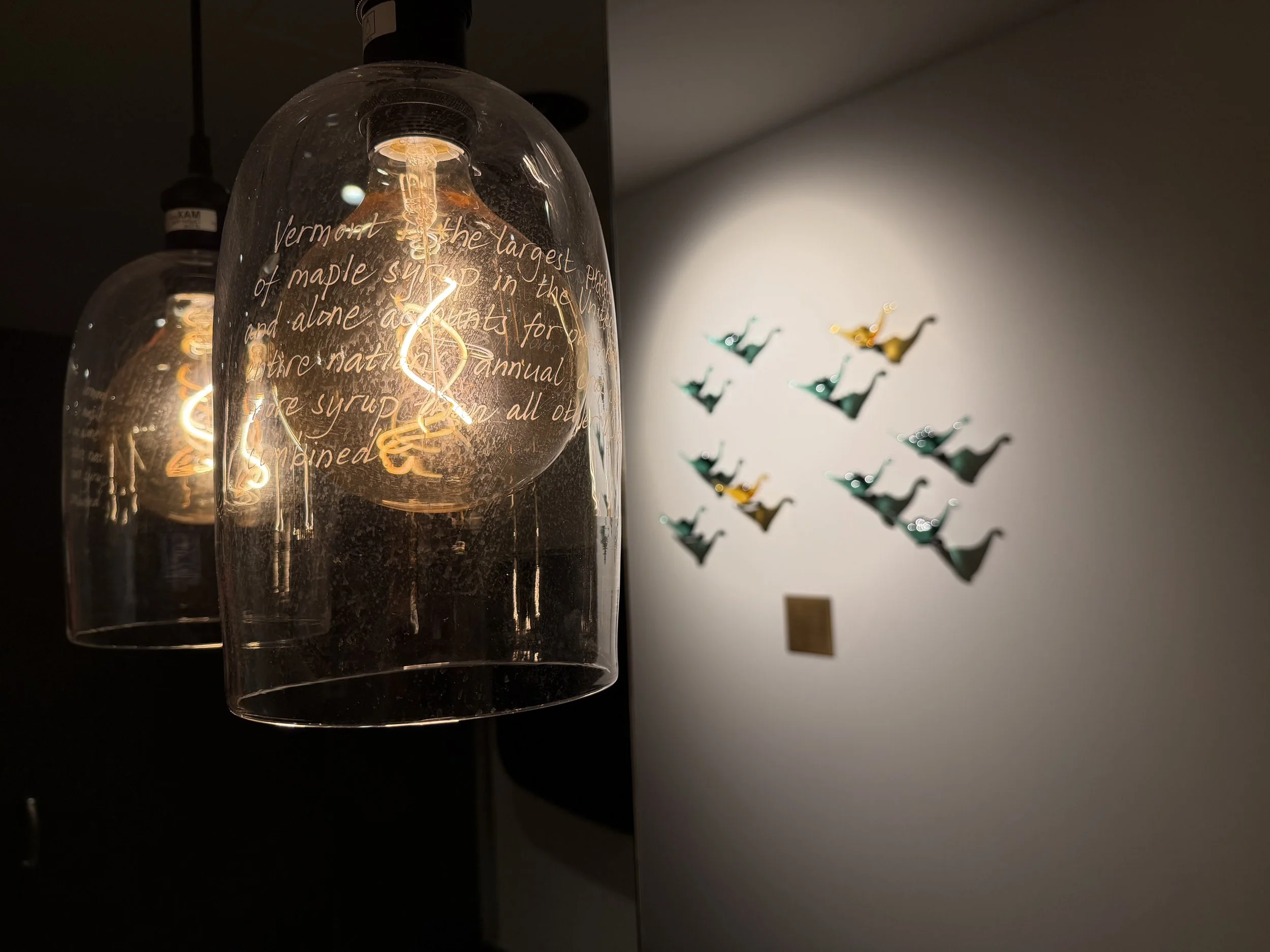 Close-up view of an illuminated filament light bulb with handwritten text on its surface, hanging from the ceiling, with a wall decorated with small colorful paper bird cutouts in the background.