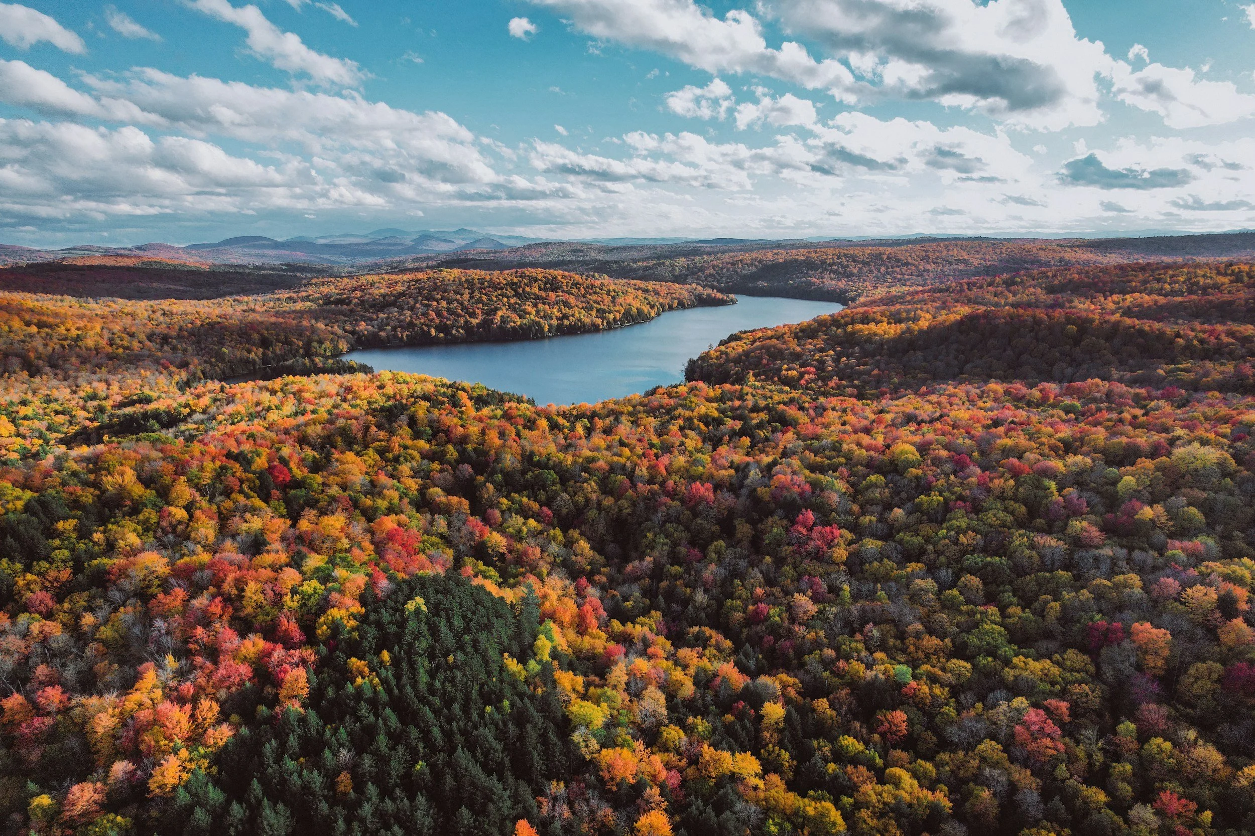 Aerial view of a large lake surrounded by colorful autumn trees with mountains in the background and a partly cloudy sky.