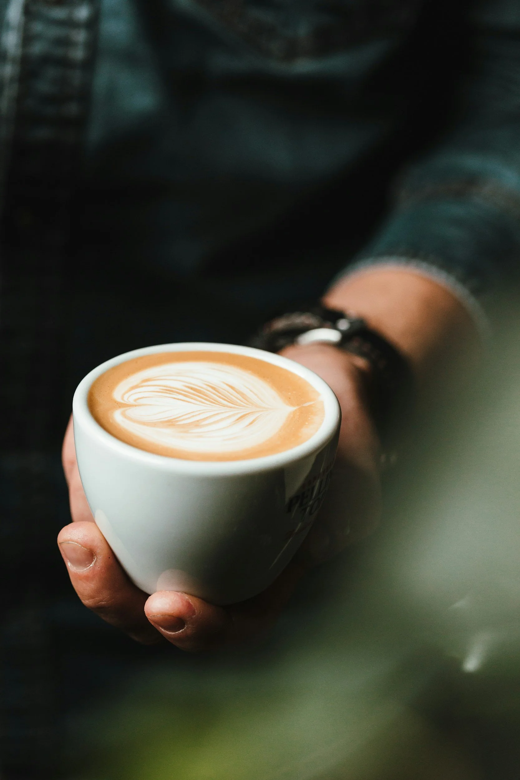 Close-up of a hand holding a white coffee mug with latte art, a heart and leaf design.