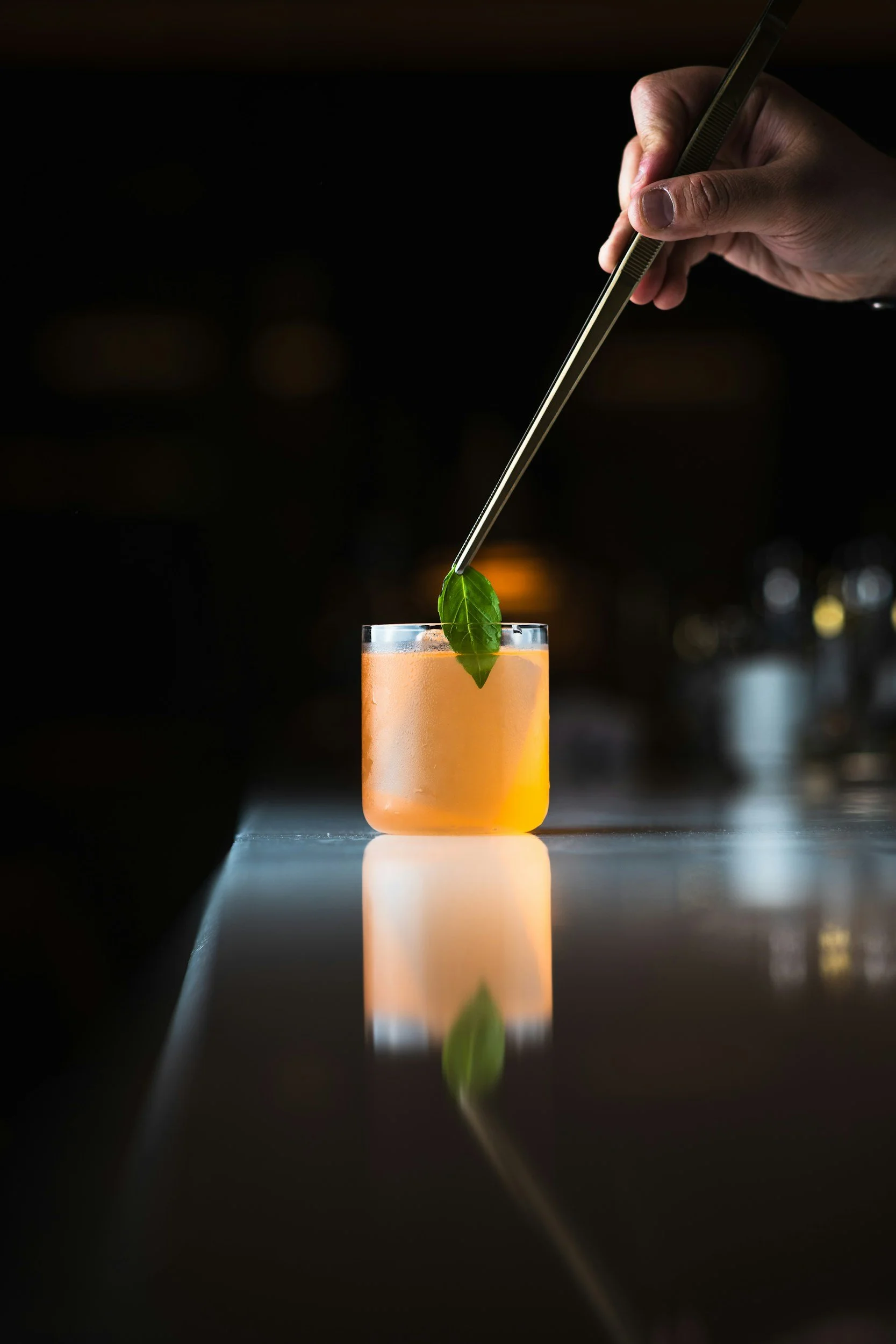 A hand holding a pair of tweezers picking up a green leaf from a tall glass filled with an orange beverage, with a reflective surface underneath.