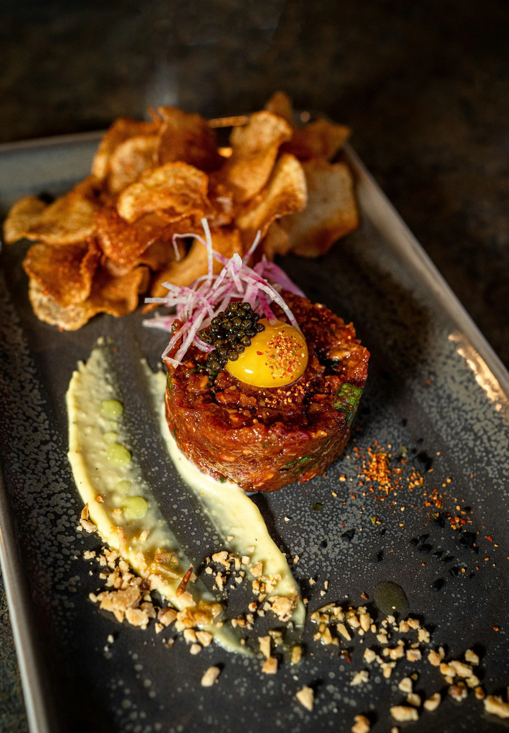 A plated gourmet appetizer featuring a piece of tuna tartare topped with a raw egg yolk and caviar, accompanied by a dollop of cream or sauce, chopped nuts, and crispy chips on a dark plate.