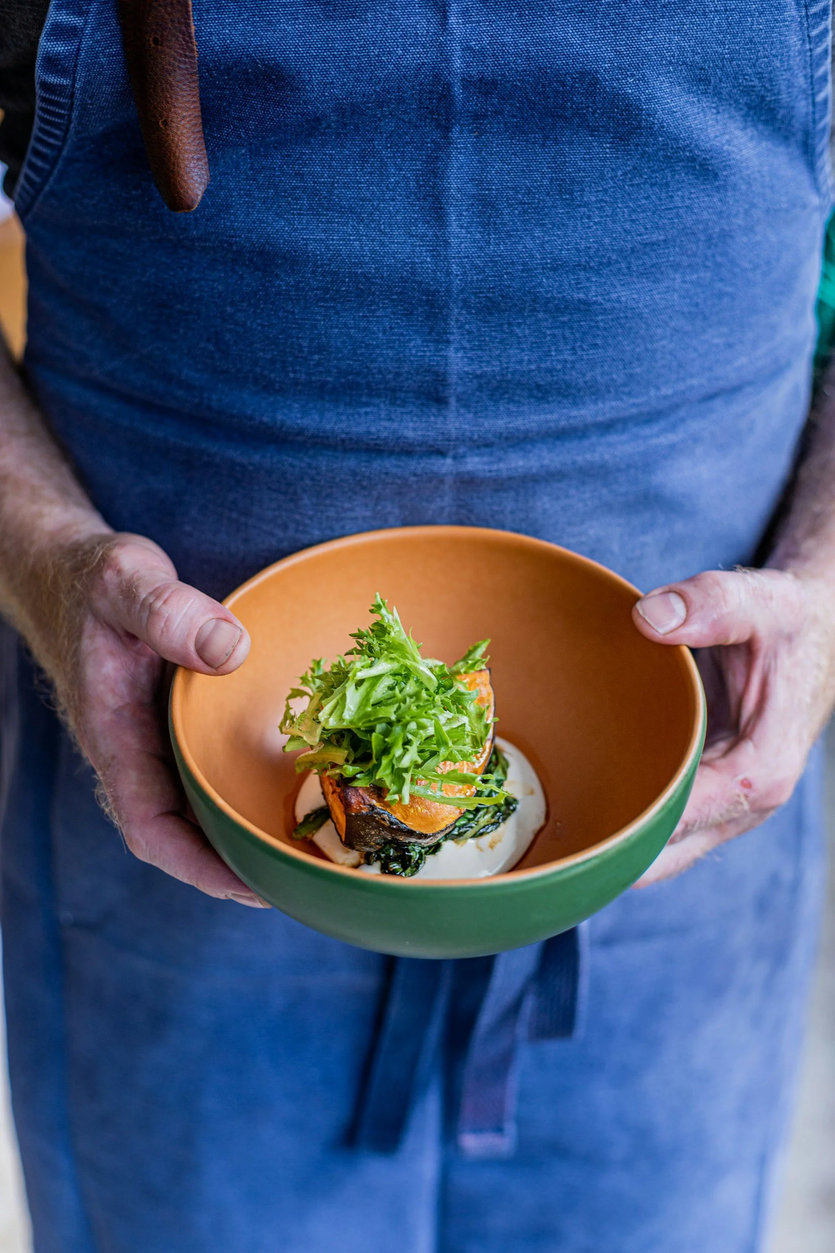 A person wearing a blue shirt holding a bowl of roasted vegetables topped with fresh greens.