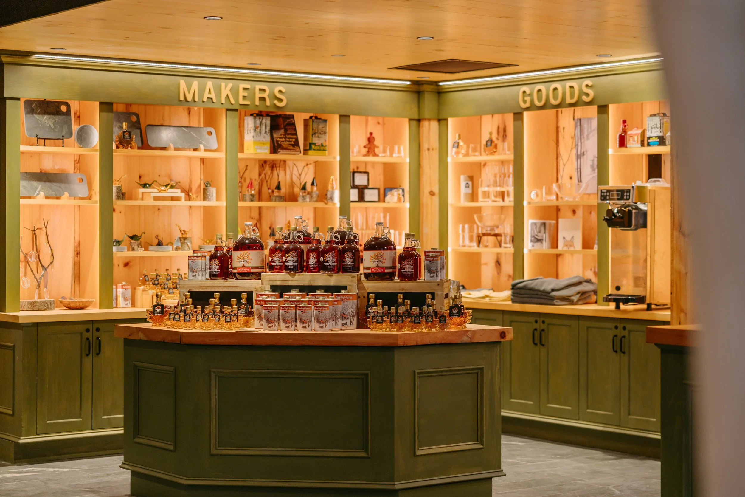 A retail display interior with shelves labeled 'MAKERS' and 'GOODS', featuring various items such as jars, bottles, books, and decorative objects, with a wooden and green color scheme.