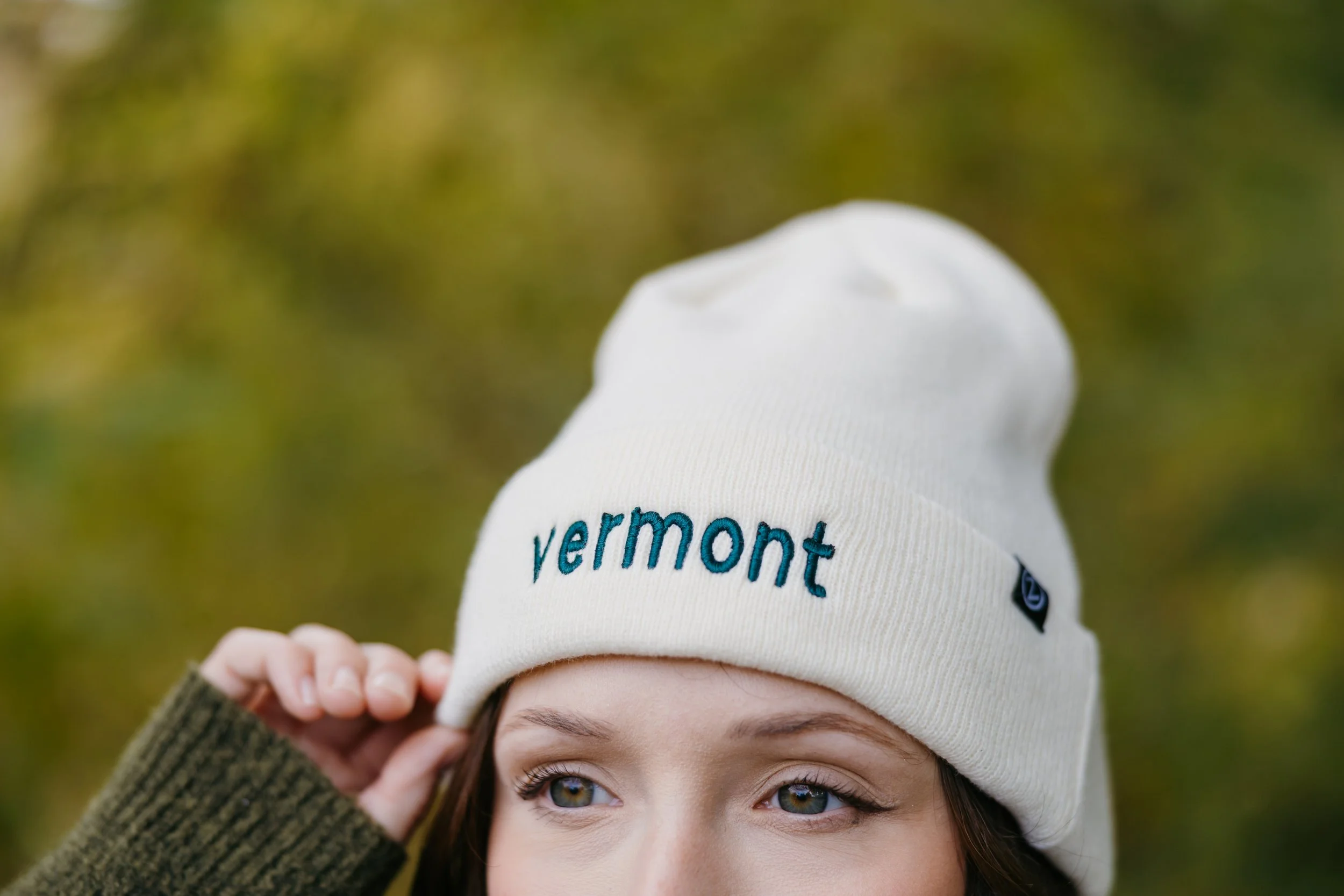 A person with blue eyes is wearing a white beanie with the word 'Vermont' embroidered on it. They are outdoors with a blurred green background.