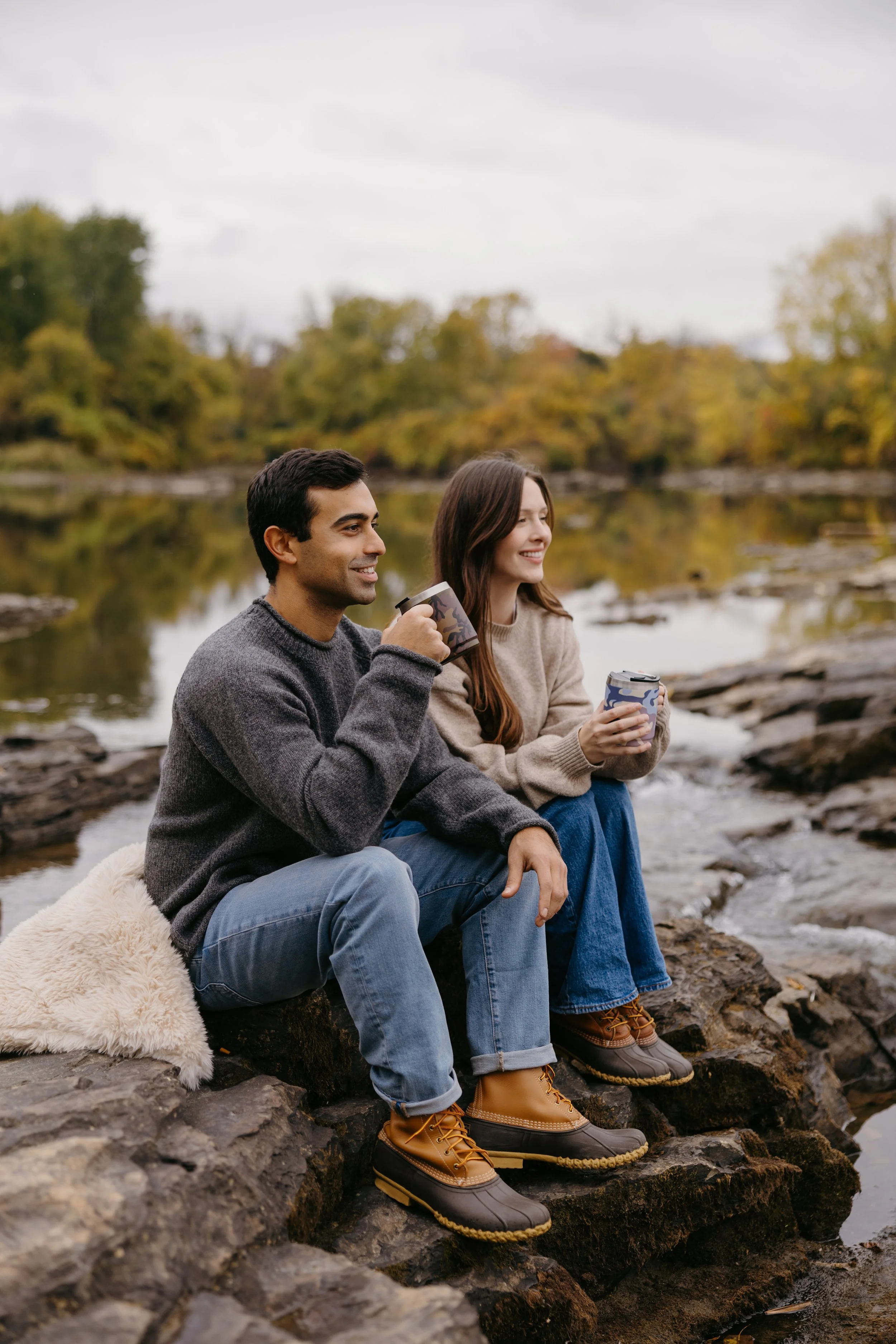A man and a woman sitting by a river during fall, holding coffee mugs, smiling, wearing warm clothing, with trees in full autumn colors in the background.
