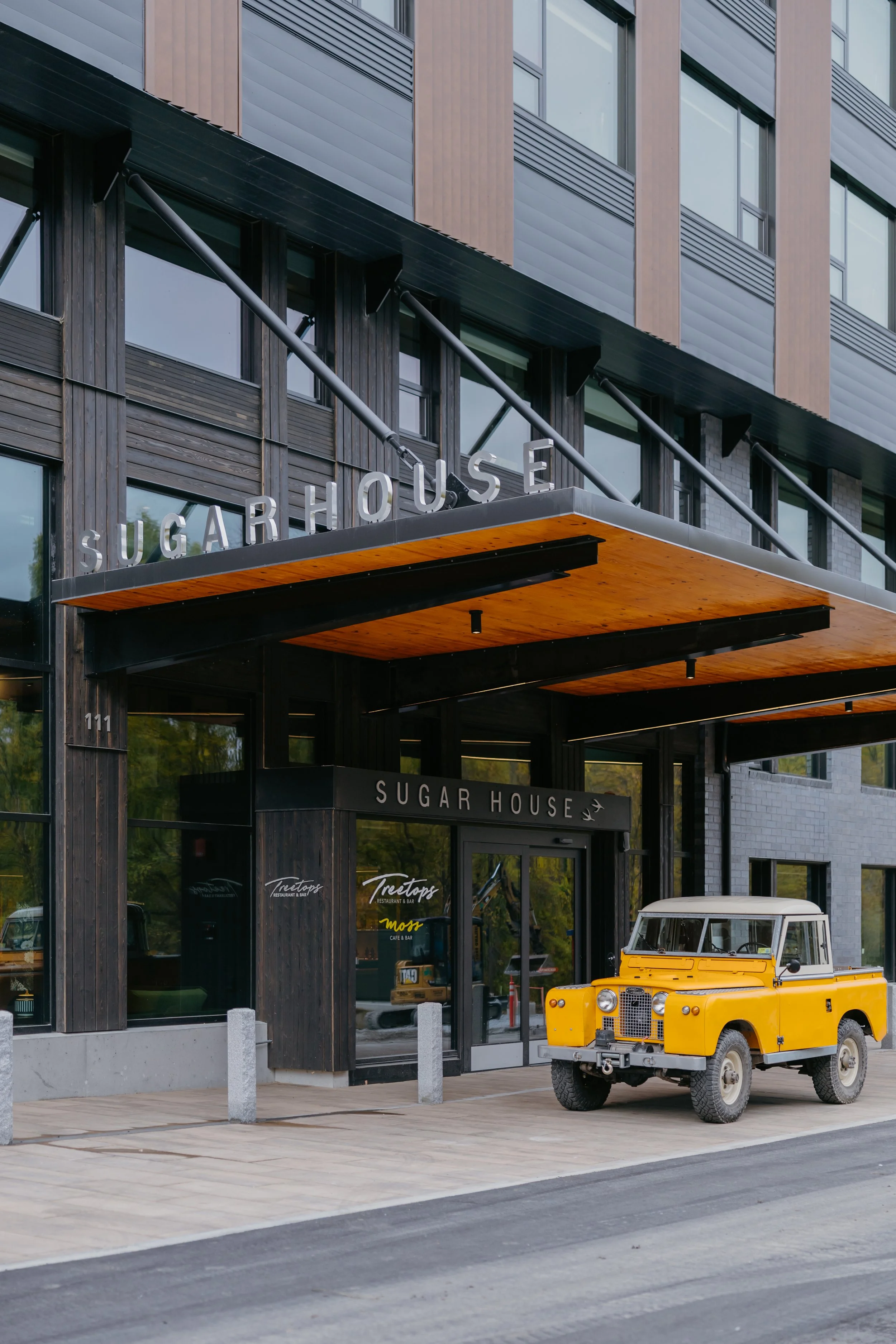 The image shows the entrance of a modern building with the sign 'Sugar House' above the doorway. There is a bright yellow vintage off-road vehicle parked in front, and a golf cart is visible on the right. The exterior features a mix of dark wood and 