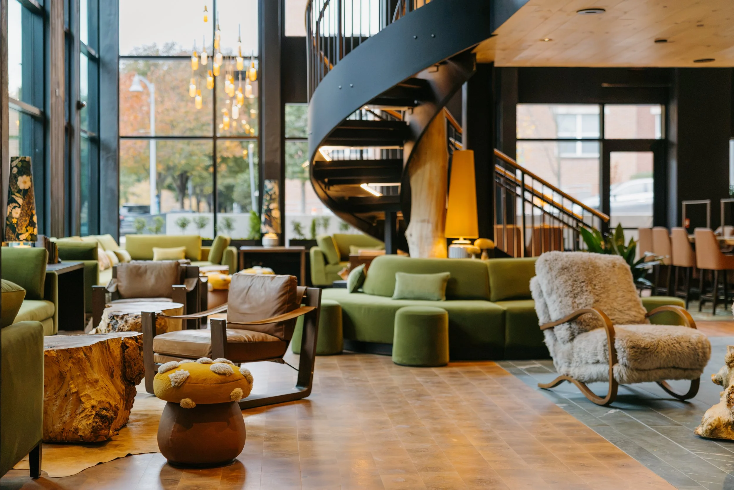 Interior of a modern hotel lobby with large glass windows, green sofas, a spiral staircase, various chairs, and decorative lighting fixtures.