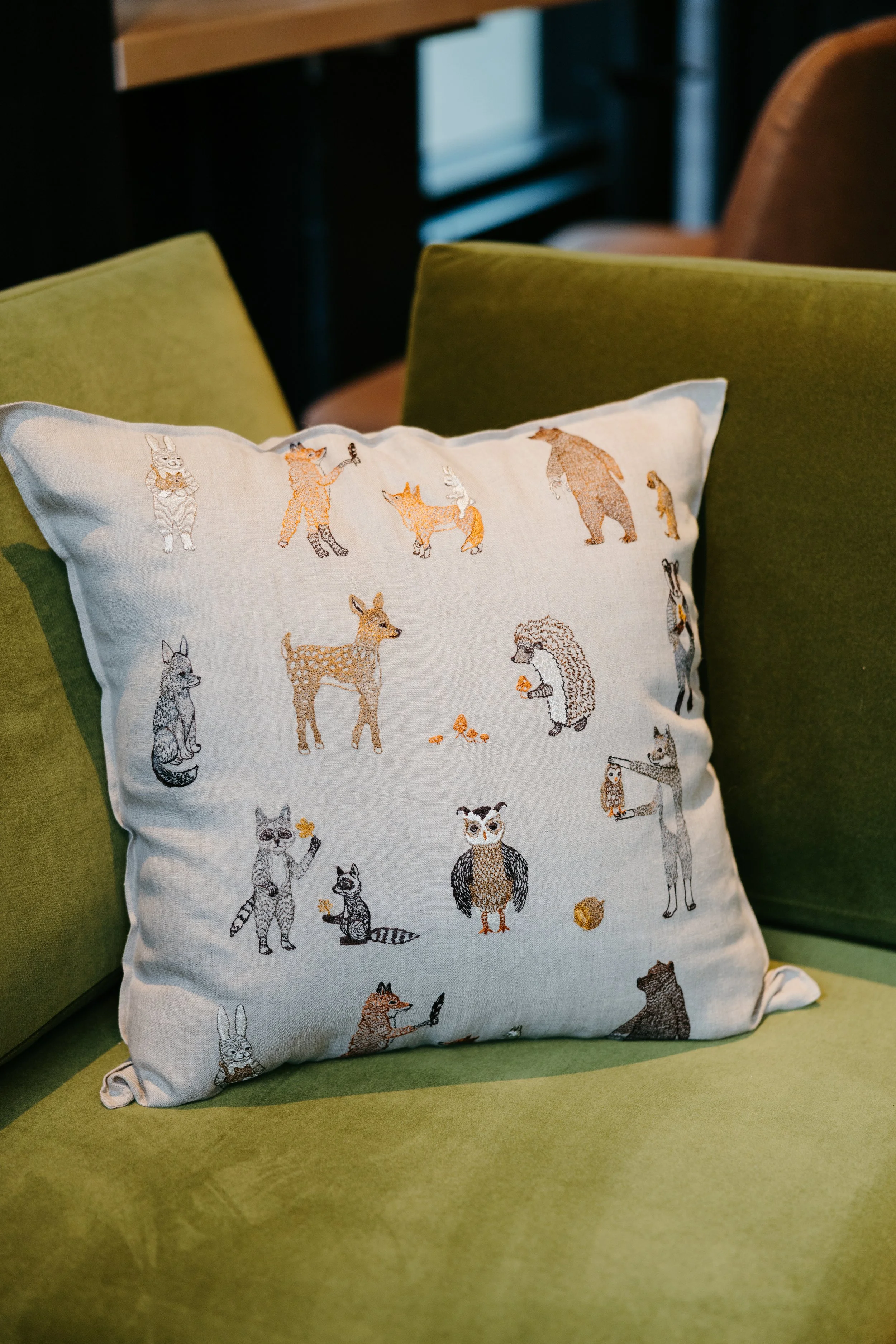 A decorative pillow with embroidered animals including a fox, bear, deer, owl, raccoon, and rabbit, resting on a green chair with another green cushion behind it.
