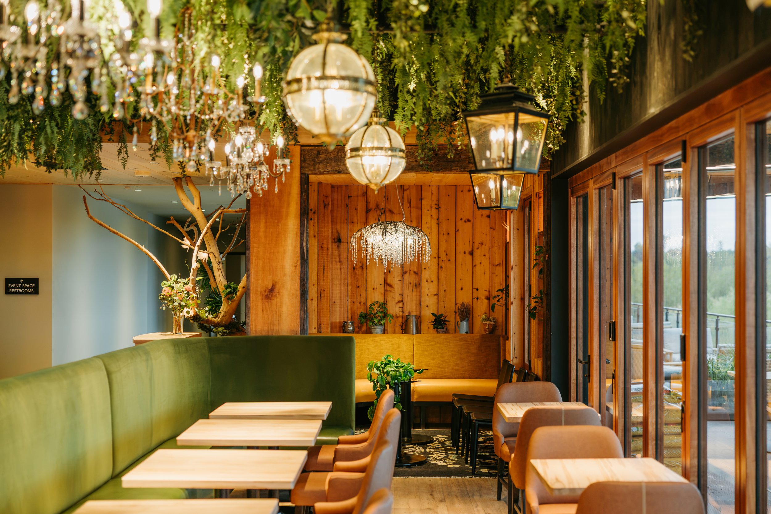 Interior of a cozy café with a green upholstered bench, wooden tables, and chairs. The ceiling is decorated with hanging greenery, crystal chandeliers, and vintage lanterns. Large windows on the right let in natural light.