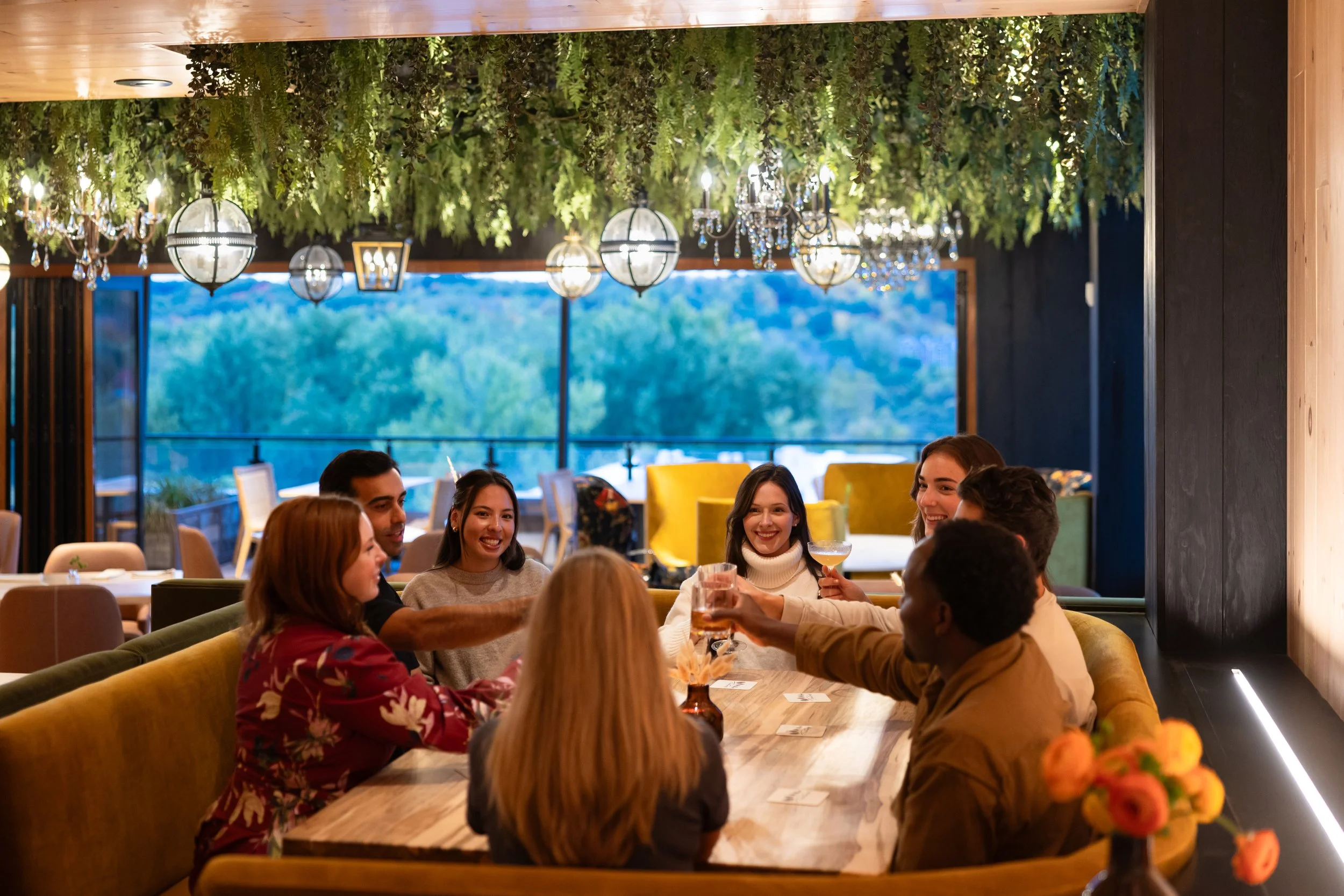 A group of people sitting at a dinner table, smiling and enjoying drinks, inside a warmly lit restaurant with chandeliers and green foliage overhead.
