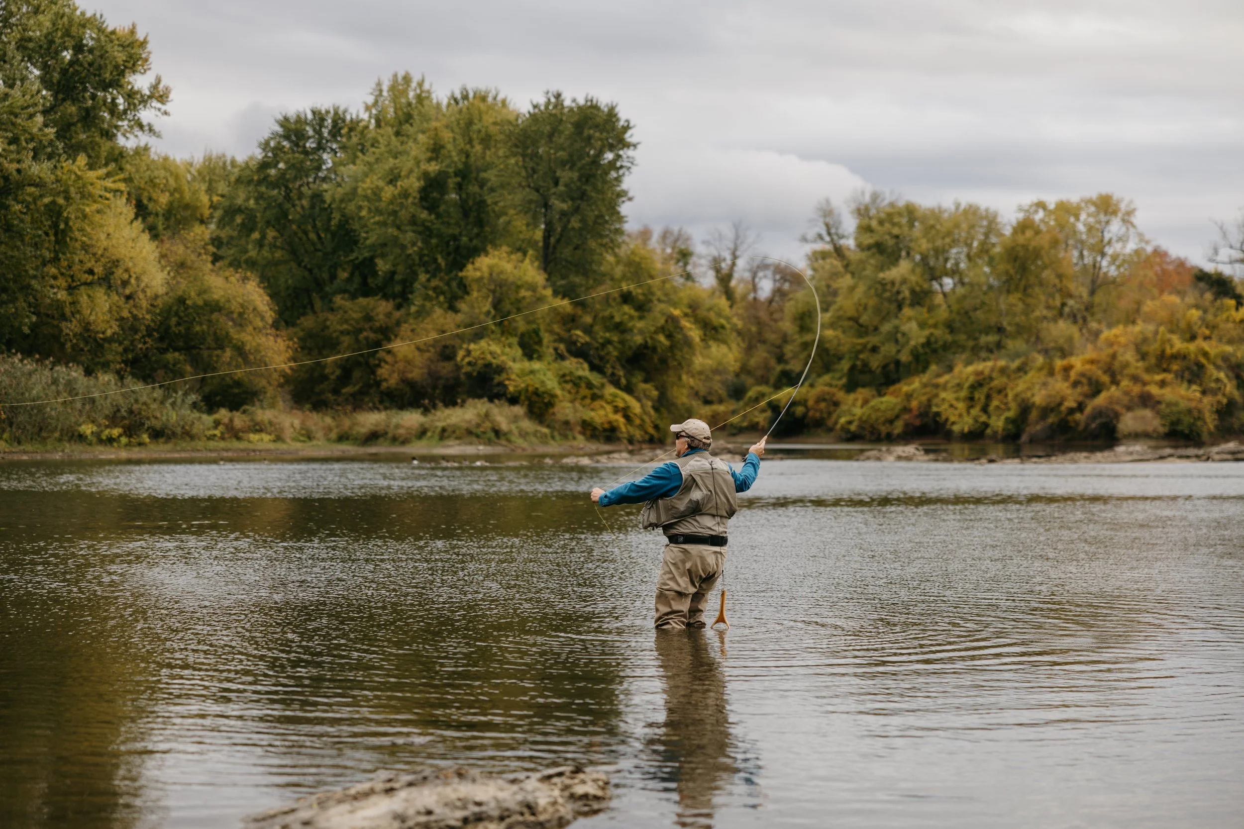 A man fly fishing in a river surrounded by trees with autumn foliage.