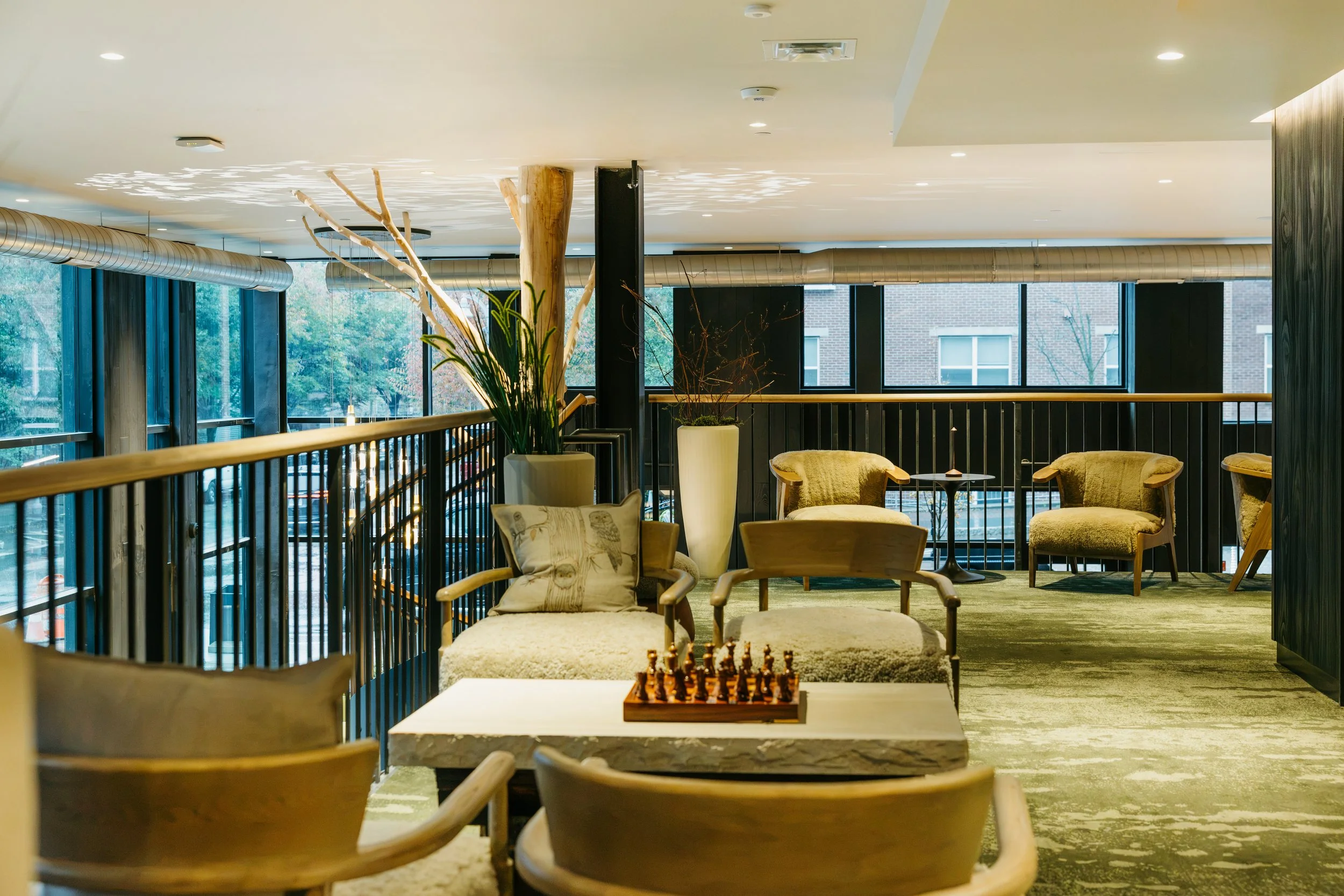 A cozy lounge area with wooden chairs, a coffee table with a chess set, large potted plants, and large windows showing trees outside.