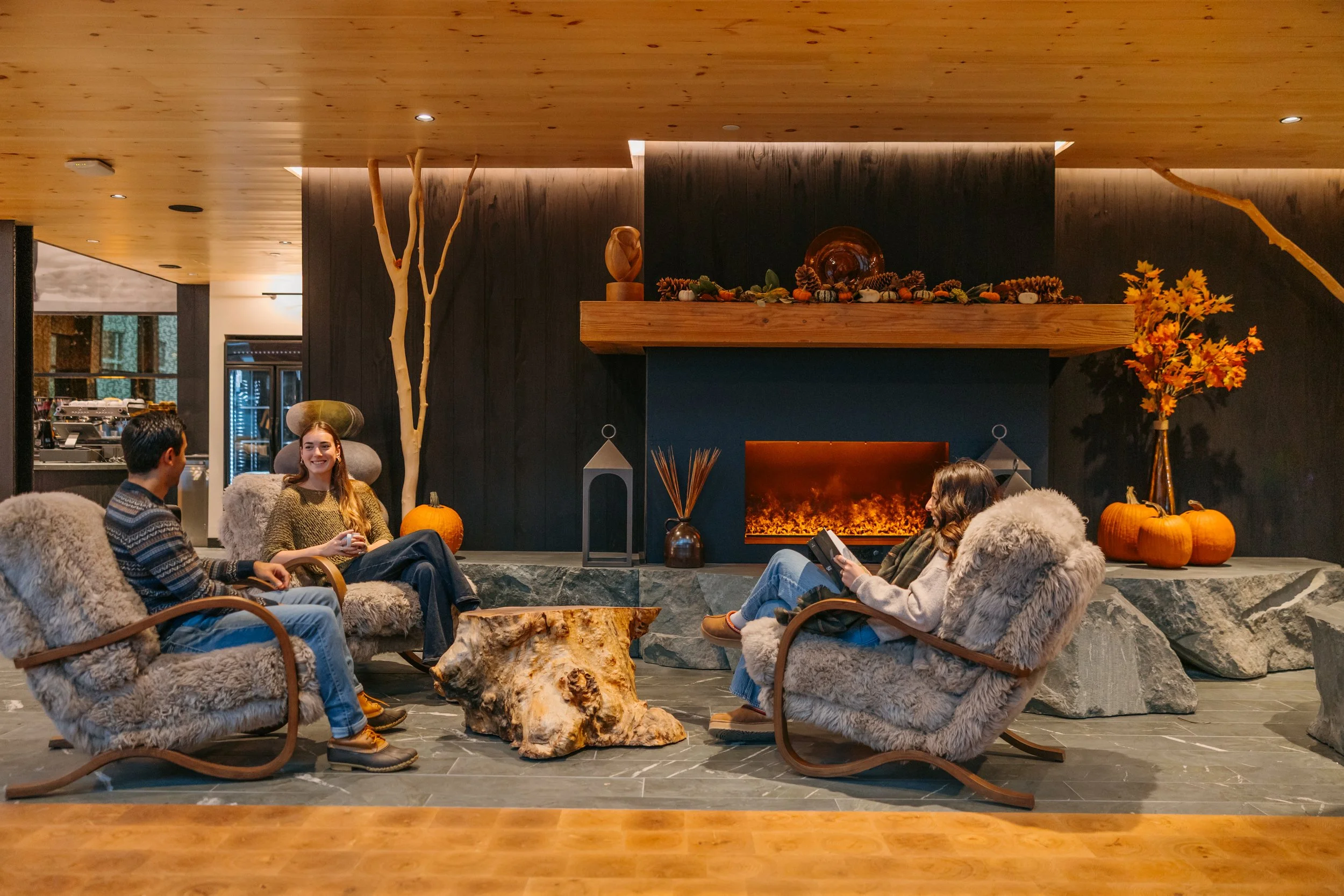 A cozy indoor scene with four people sitting in plush, furry chairs arranged in a semicircle in front of a fireplace. The fireplace is decorated with pumpkins, pine cones, and autumn leaves, indicating a fall or Thanksgiving theme. The room has wood-