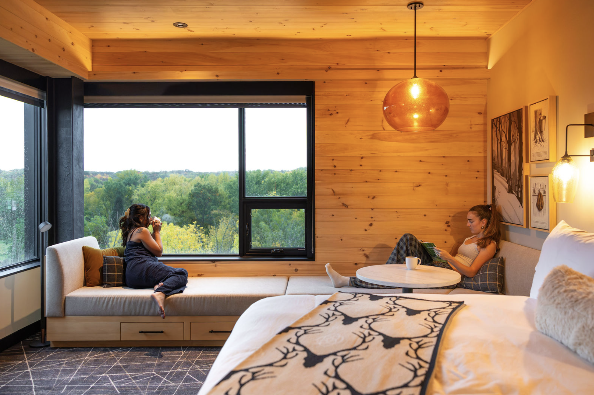 Two women relaxing inside a cozy room with wooden walls and large windows overlooking a green, wooded landscape. One woman is sitting on a white built-in sofa, drinking from a mug, while the other is reclining on a separate couch reading a book.