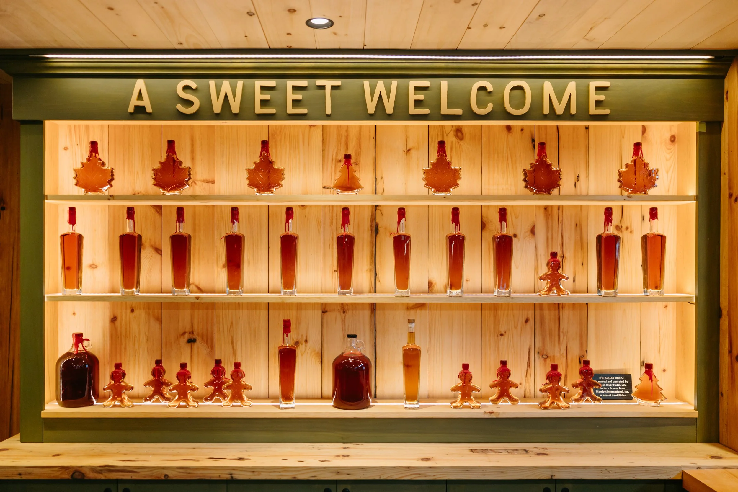 Wooden display shelf with bottles of syrup arranged in rows under a sign that reads "A Sweet Welcome".