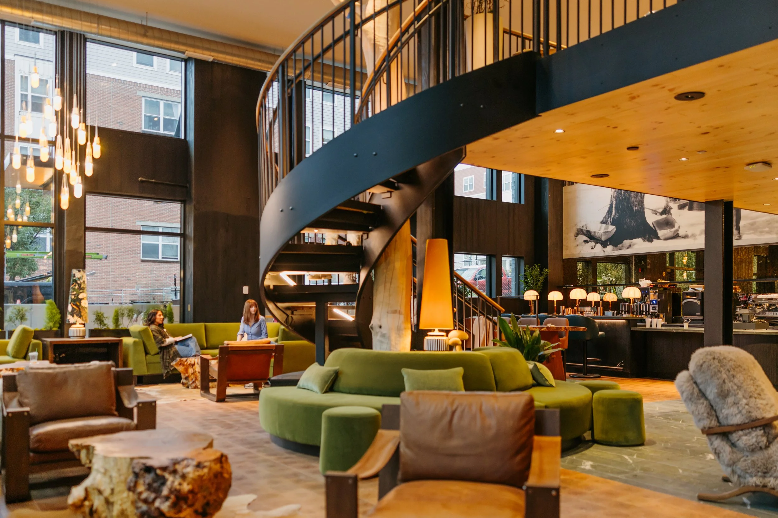 Interior of a modern hotel lobby with curved staircase, large windows, green seating, and warm lighting.