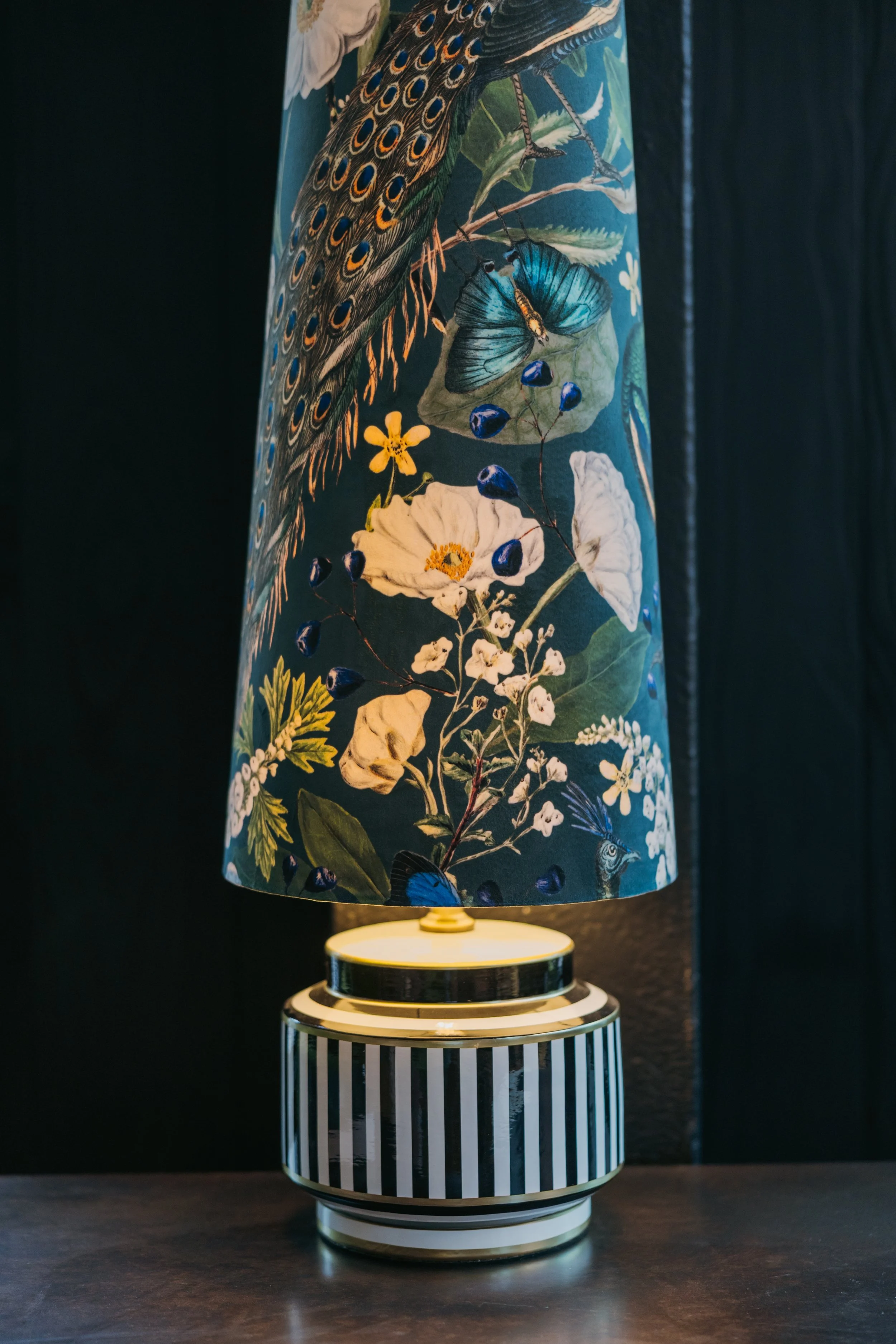A decorative table lamp with a floral and butterfly design on the lampshade, sitting on a dark surface against a black background.