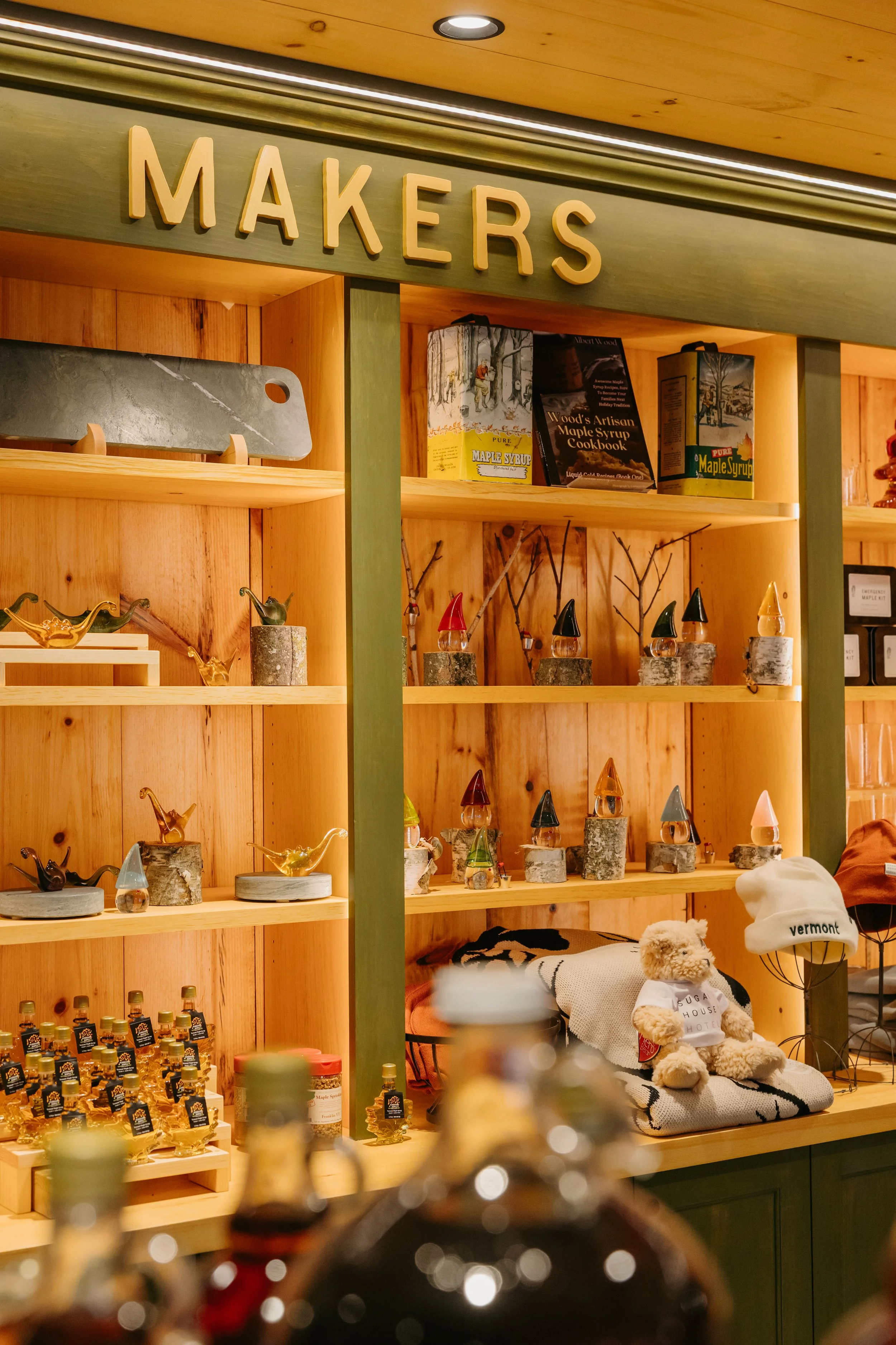 Wooden shelves displaying various jars and bottles of maple syrup and maple-related products, alongside decorative items including a stuffed teddy bear with a Vermont hat.