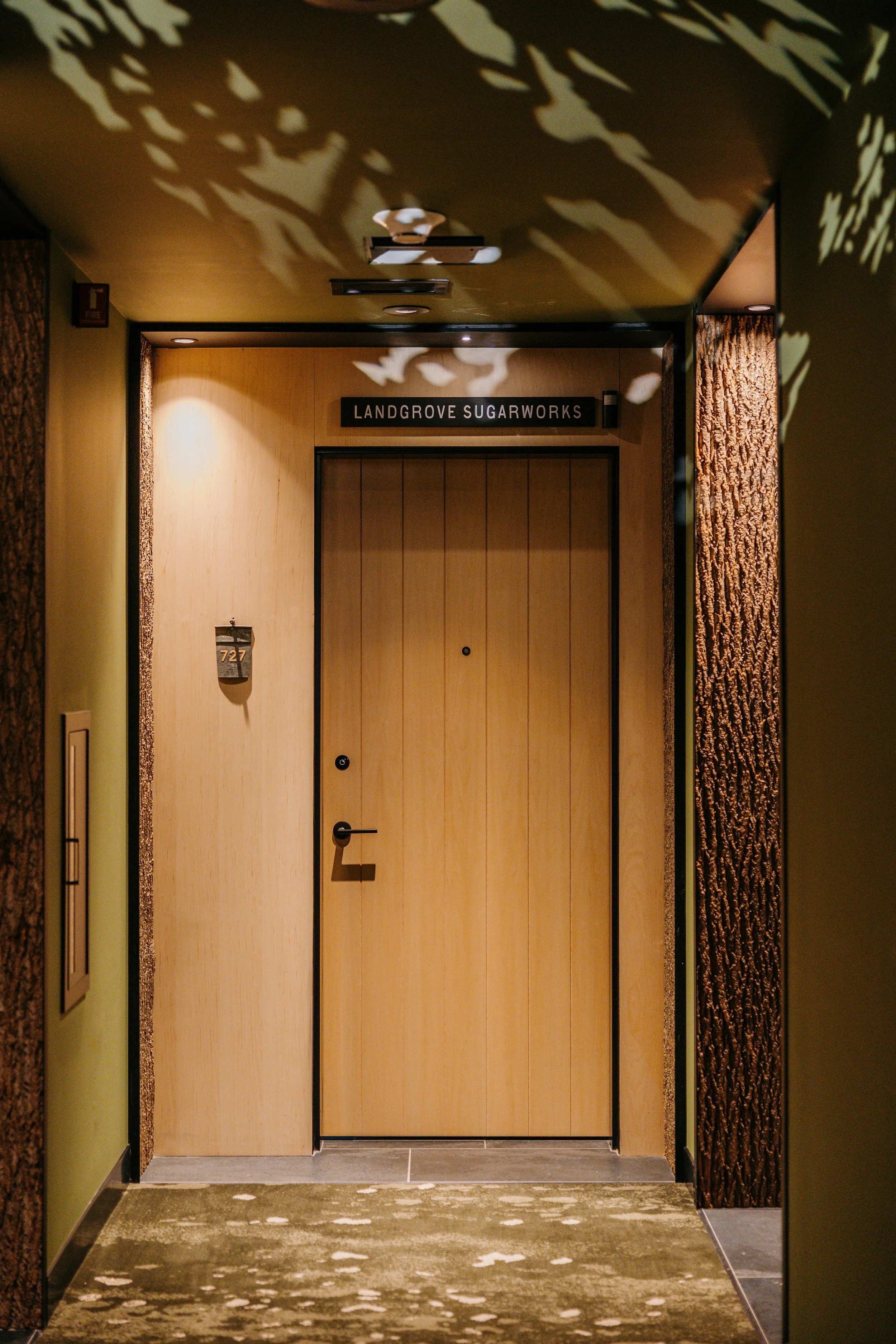 Elevator door in a hotel with the sign 'Landgrove Sugarworks' above it, surrounded by textured wood and green walls, with decorative leaf shadows on the ceiling.