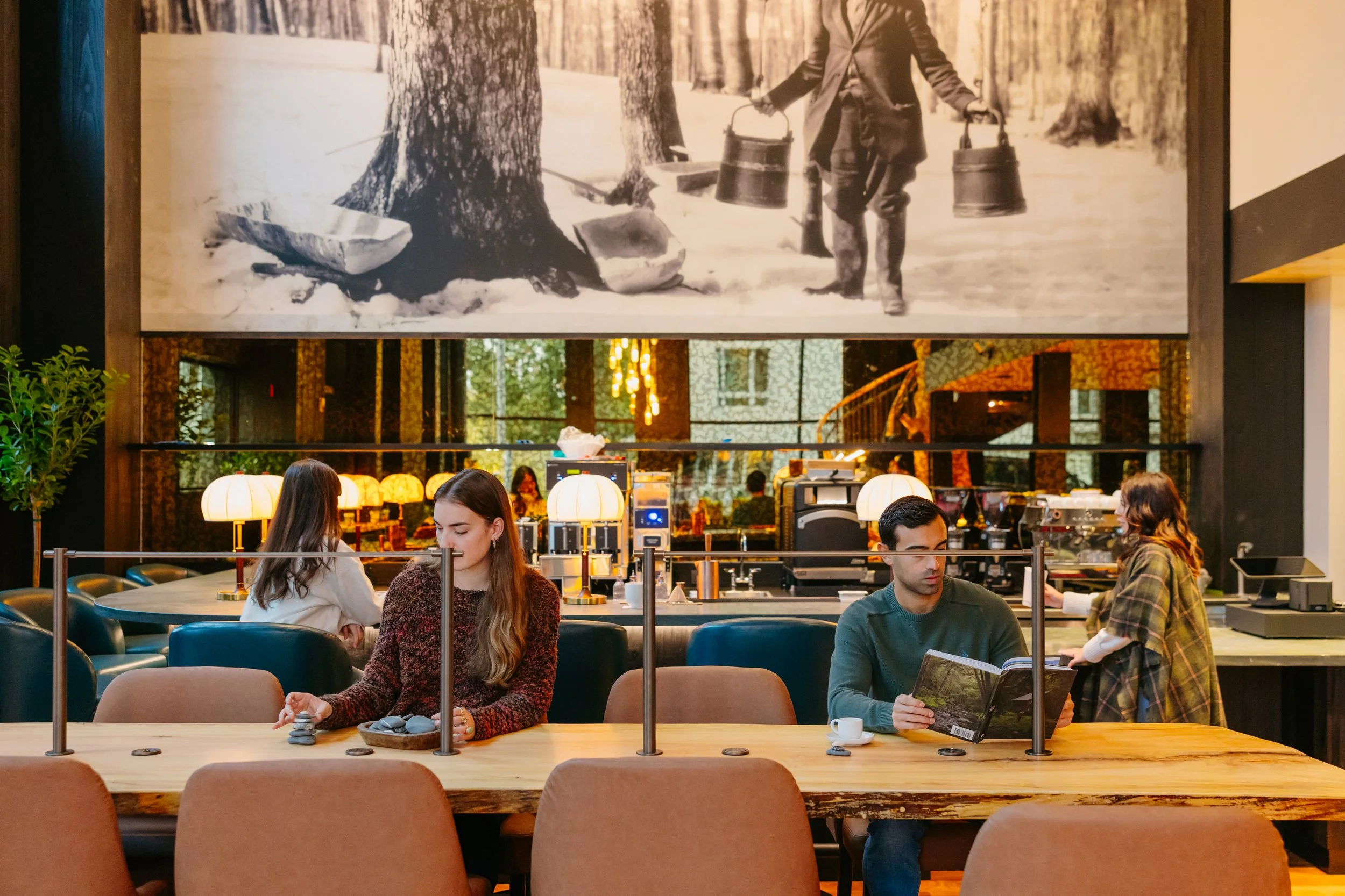 A busy cafe with diners, including two women and a man, sitting at a counter with a barista, colorful lamps, and large artwork of a person with buckets in a forest hanging on the wall.