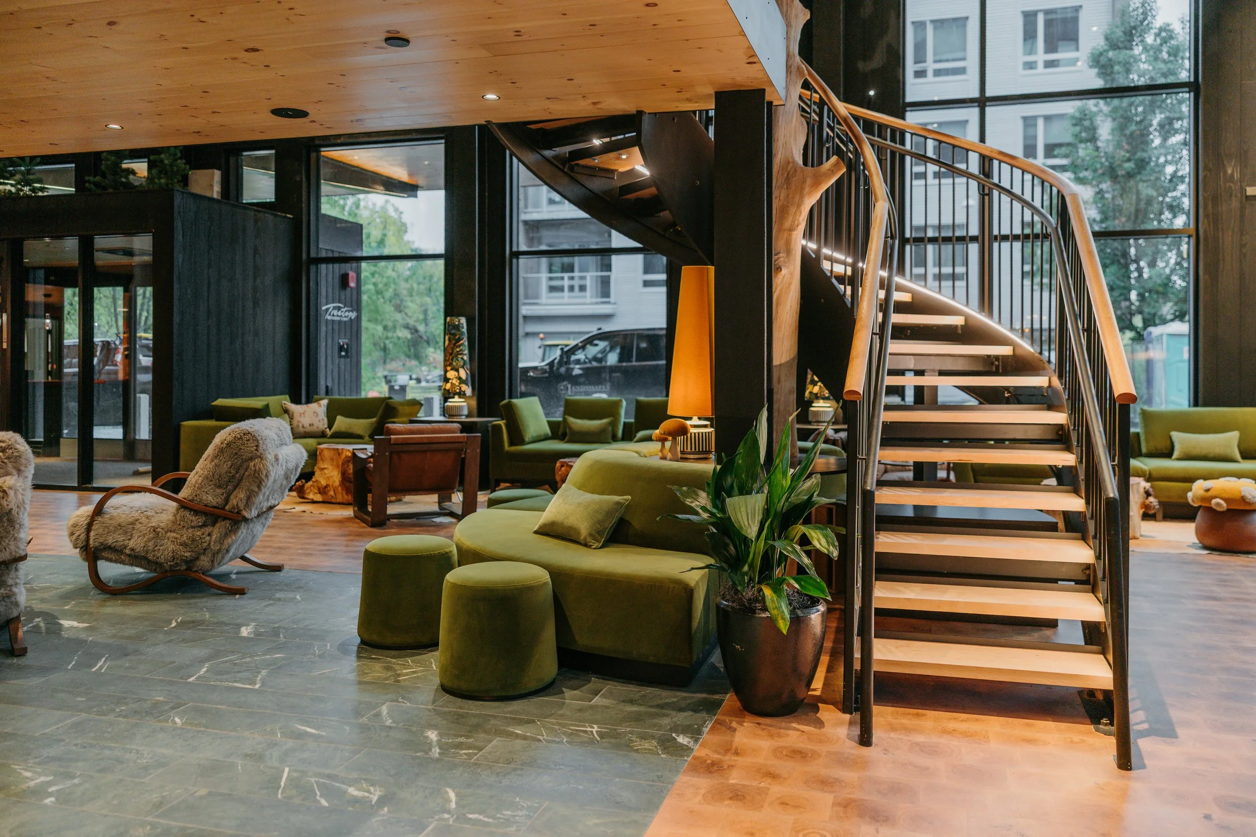 Interior view of a modern building with a curved staircase, green lounge chairs, a wooden ceiling, large windows, and decorative plants.
