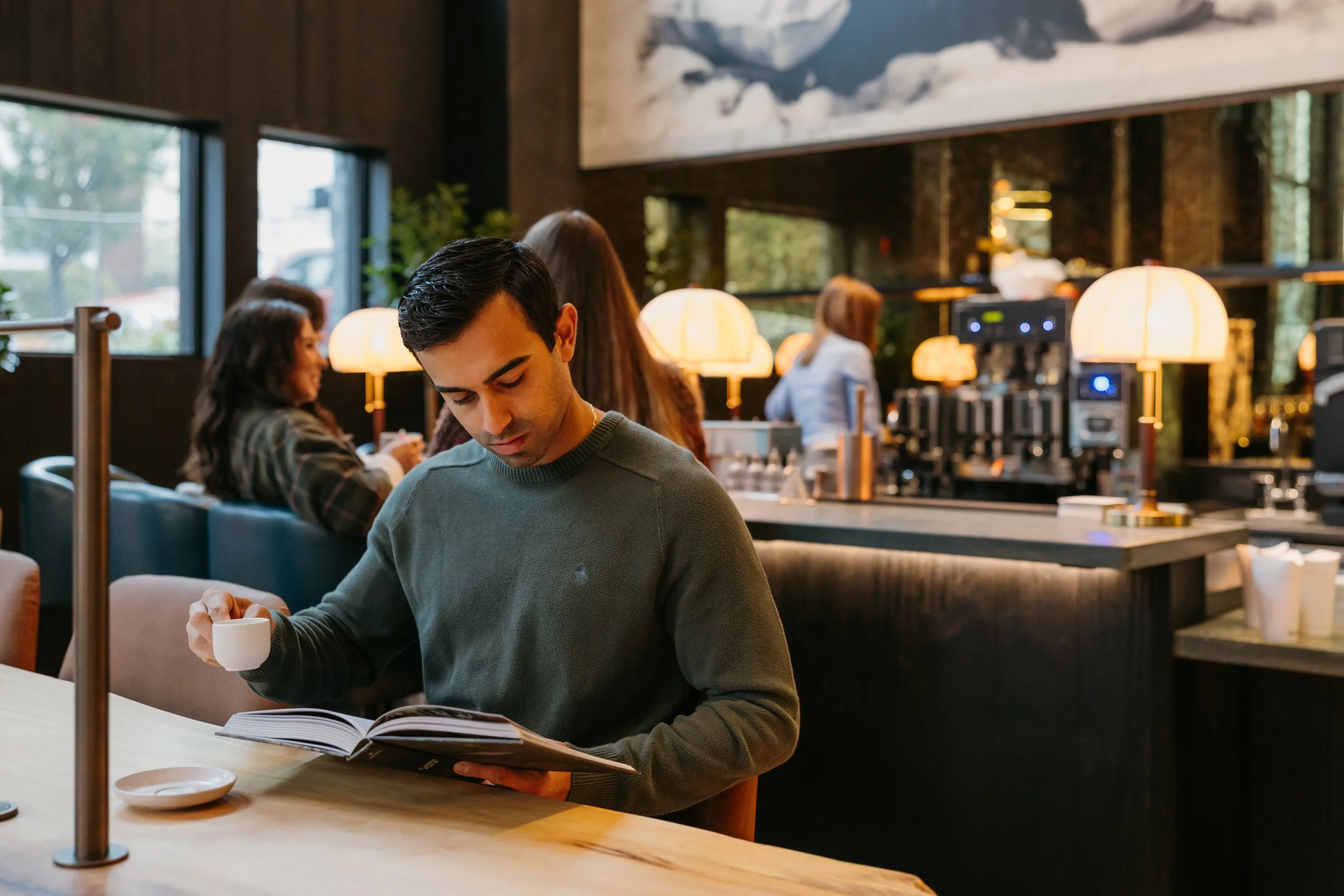 A young man with dark hair and a green sweater sitting at a wooden table in a cozy restaurant or cafe, reading a book and holding a small cup. The background features warm lighting, other patrons, large windows, and artwork on the walls.
