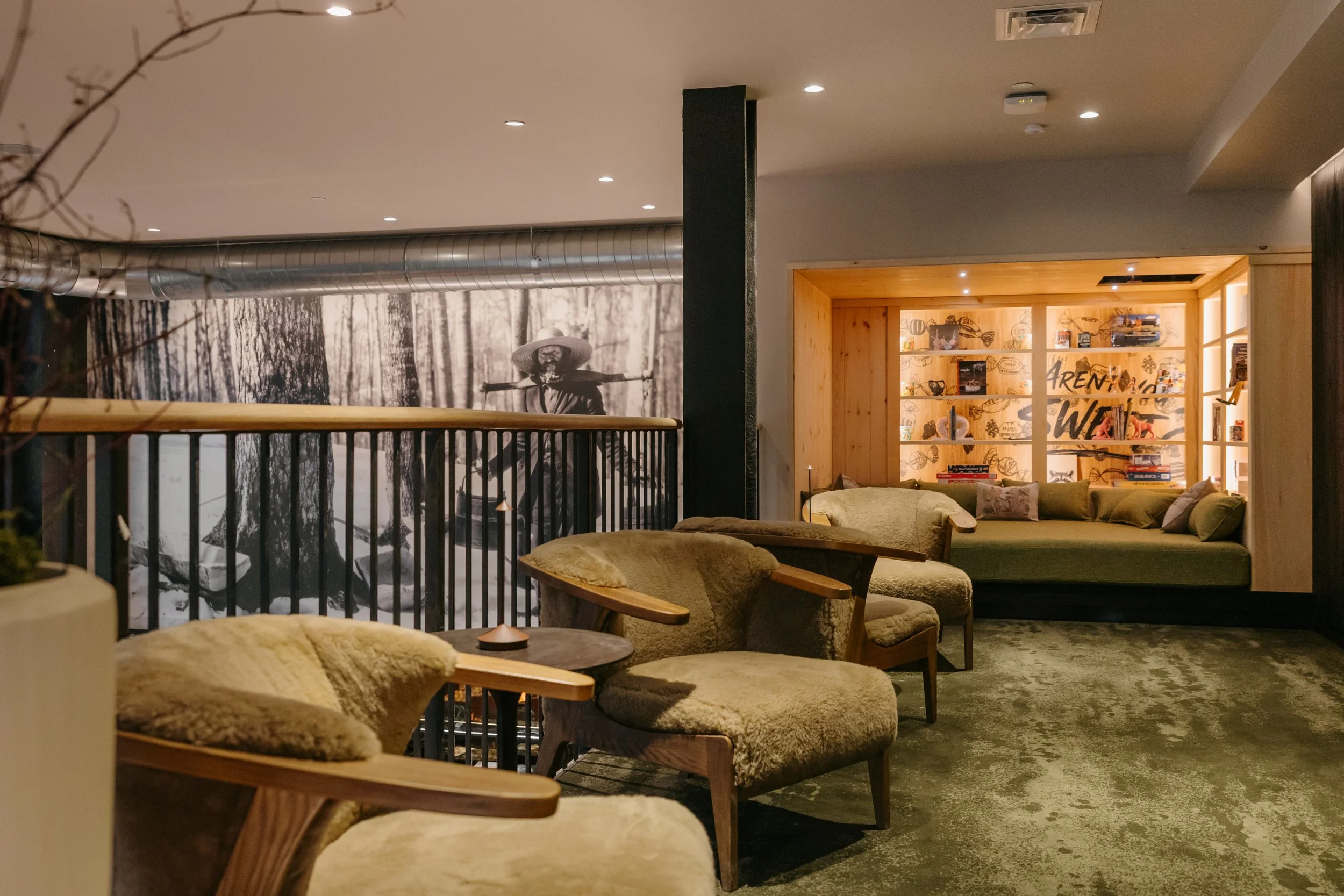 Interior of a cozy living space with beige and wooden furniture, a black-and-white forest mural on one wall, and a bookshelf in the background.