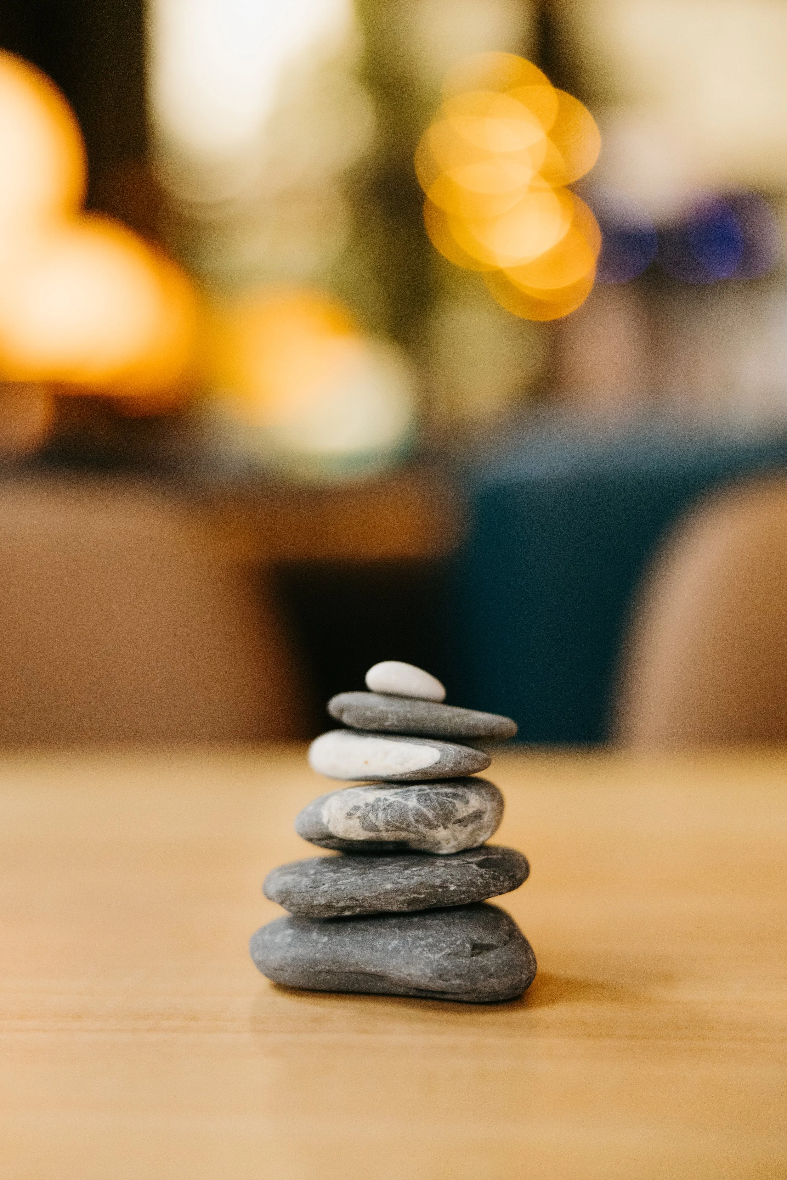 A stack of smooth gray stones arranged in a balanced tower on a wooden surface with blurry warm-colored lights in the background.