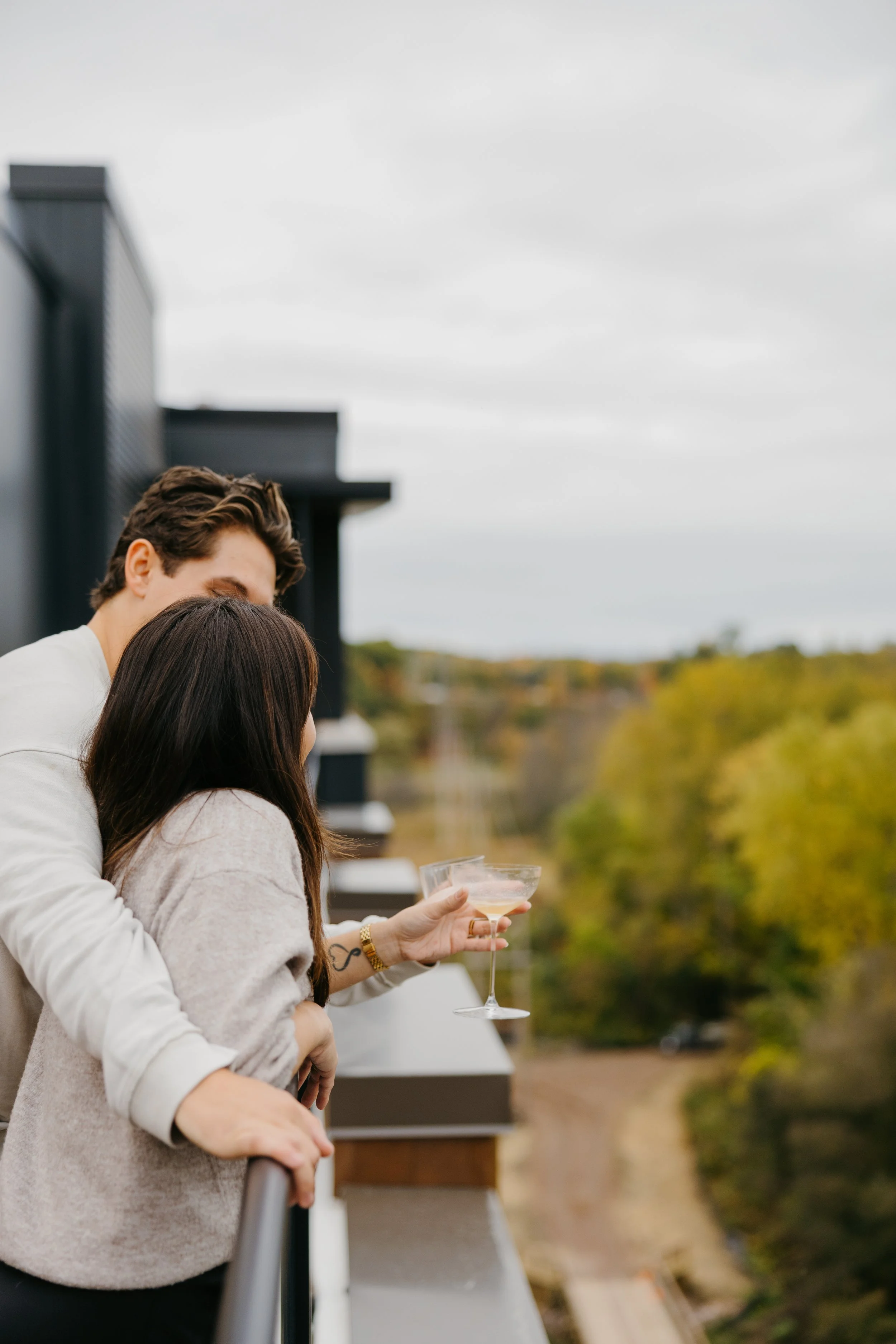 Two people, a man and a woman, standing outdoors on a balcony or rooftop, close together with the woman holding a wine glass, and cityscape and trees in the background.