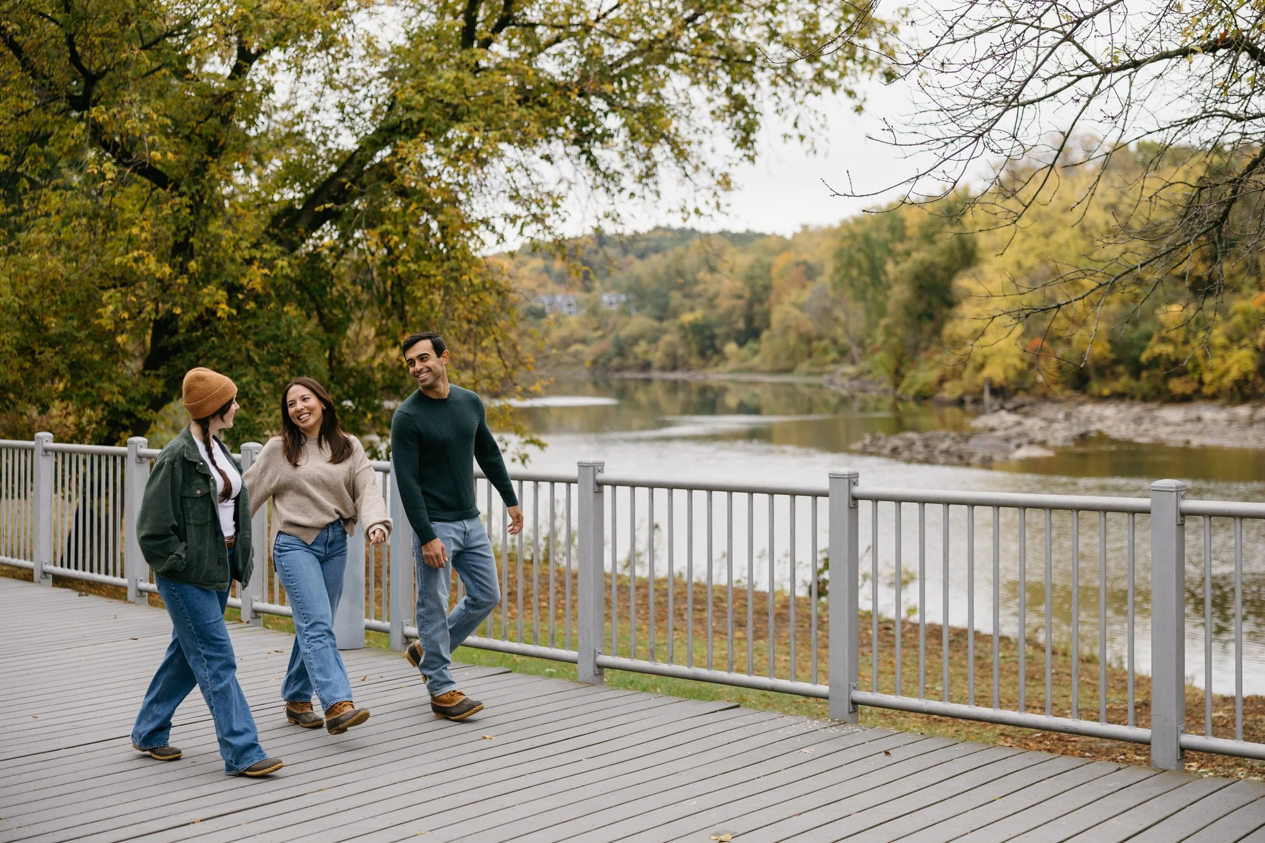 Three friends walking together on a gray wooden bridge by a river, surrounded by trees with autumn foliage.