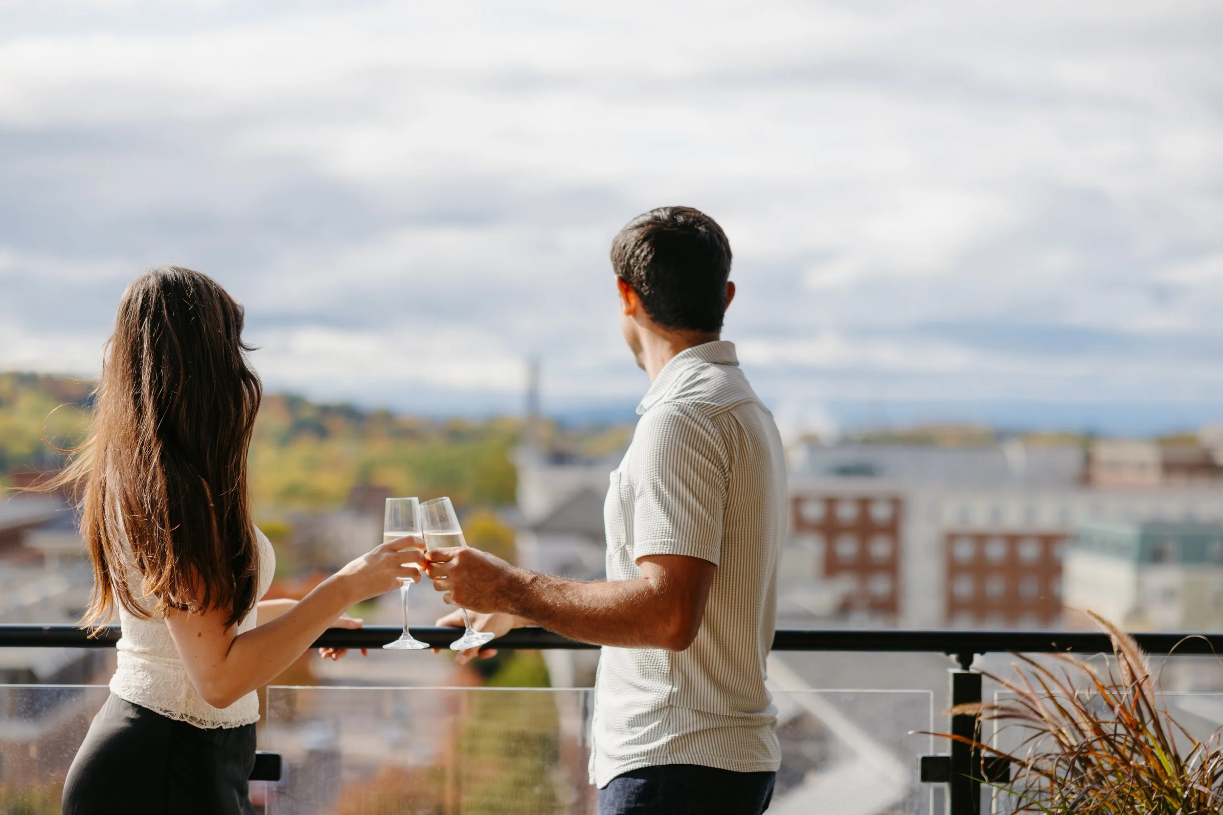 A man and woman on a balcony holding glasses of champagne, with a cityscape and cloudy sky in the background.