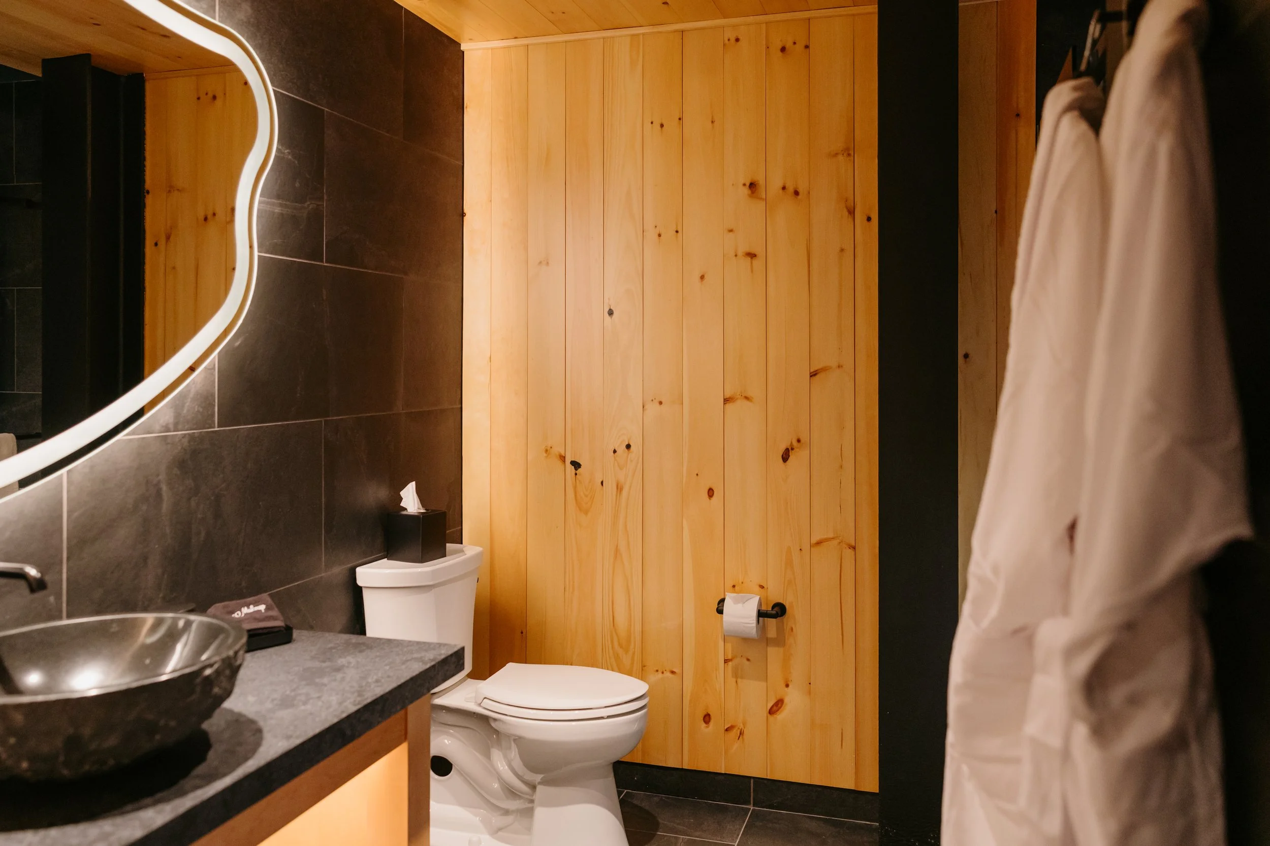 A bathroom with black tiled floor and walls, a vanity with a dark countertop, a round vessel sink, and a mirror. There is a wood-paneled wall and a toilet with a roll of toilet paper on a black holder. Part of a person wearing a beige shirt is visibl