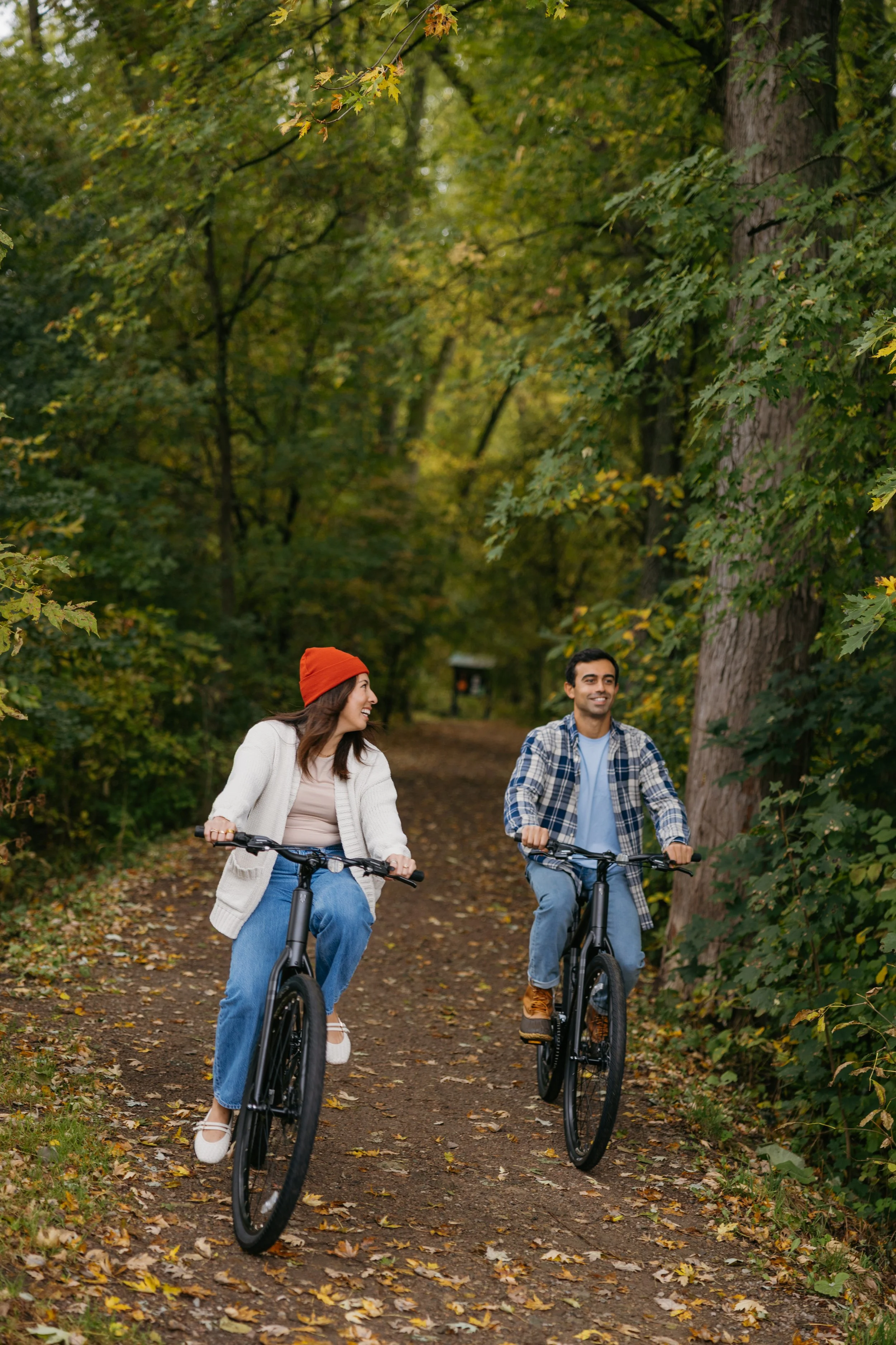 Two people riding bicycles on a forest trail with green and yellow autumn leaves, smiling and engaging with each other.