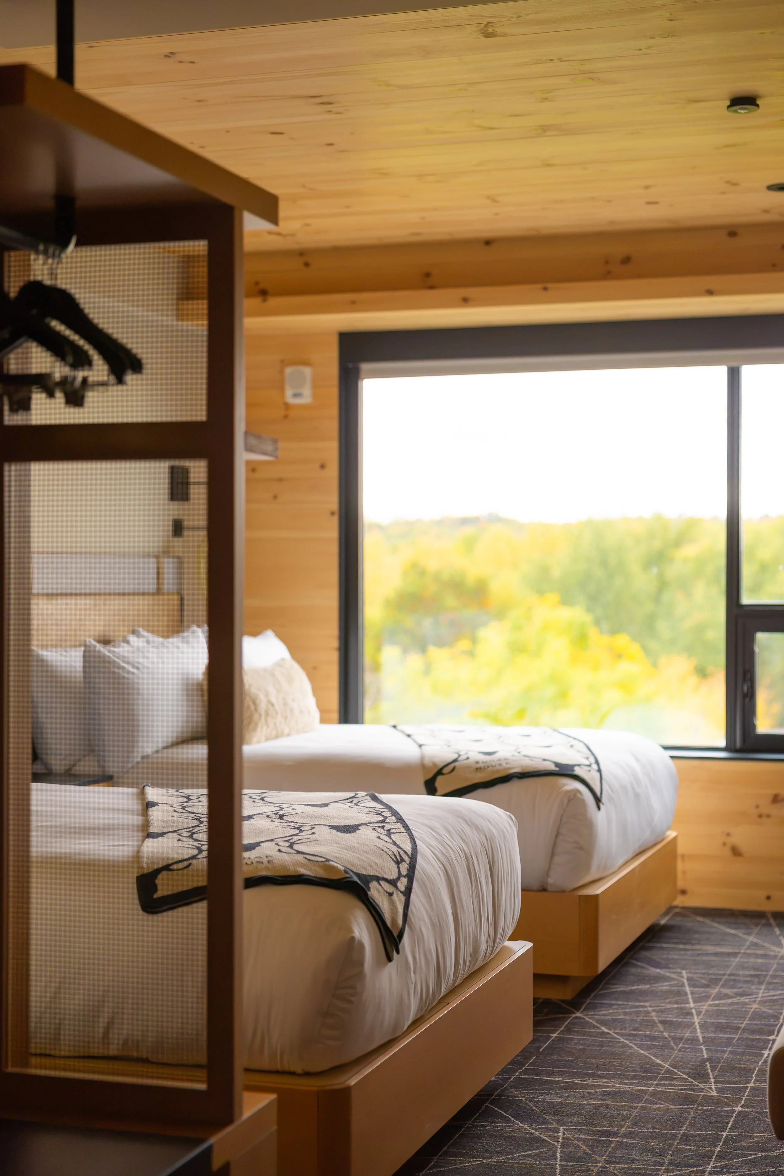 Two neatly made beds with white linens and decorative black and beige blankets in a cozy wooden-room hotel or cabin with large windows showing a scenic view of trees in autumn.