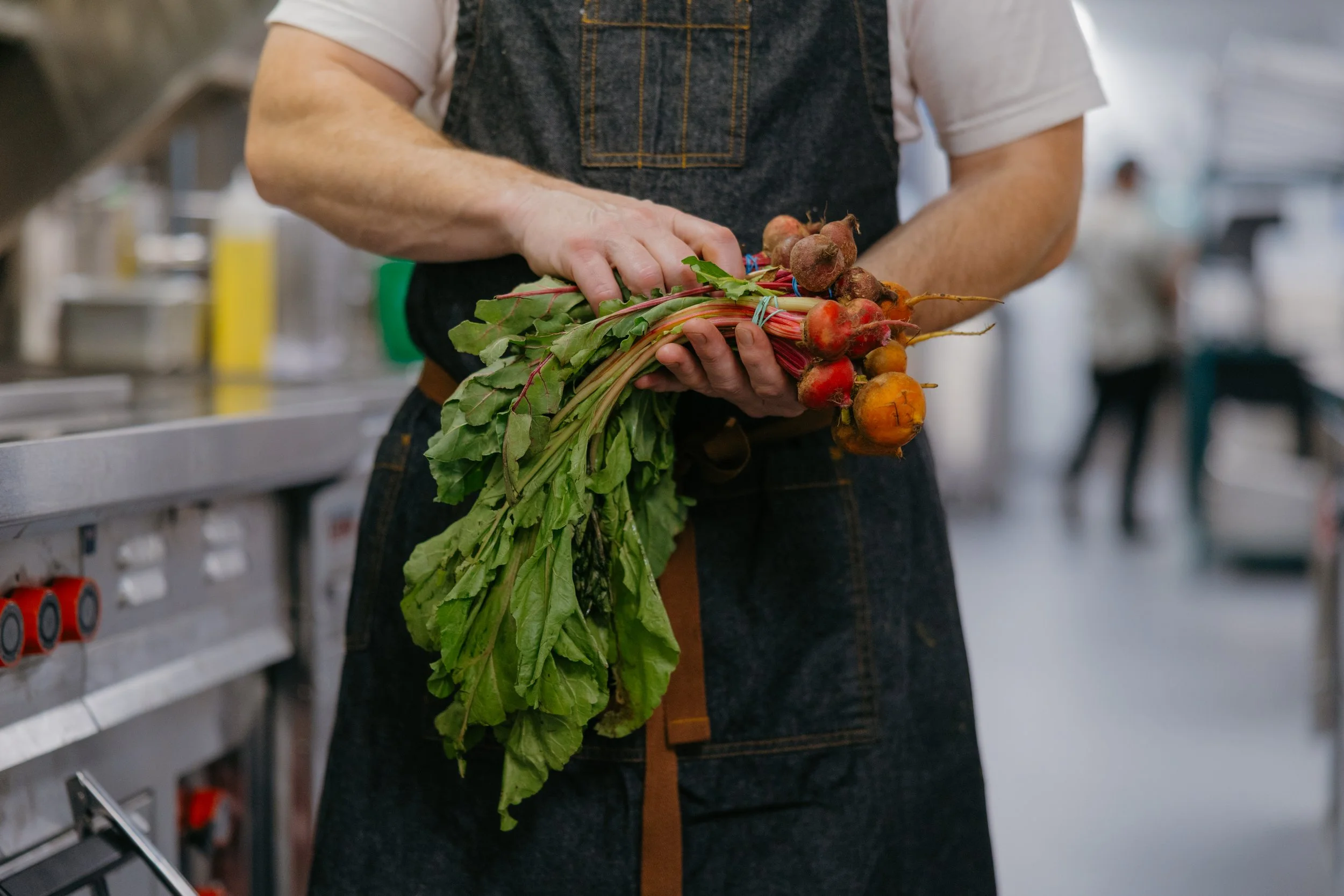Person holding fresh carrots with green leaves in a kitchen or food preparation area.