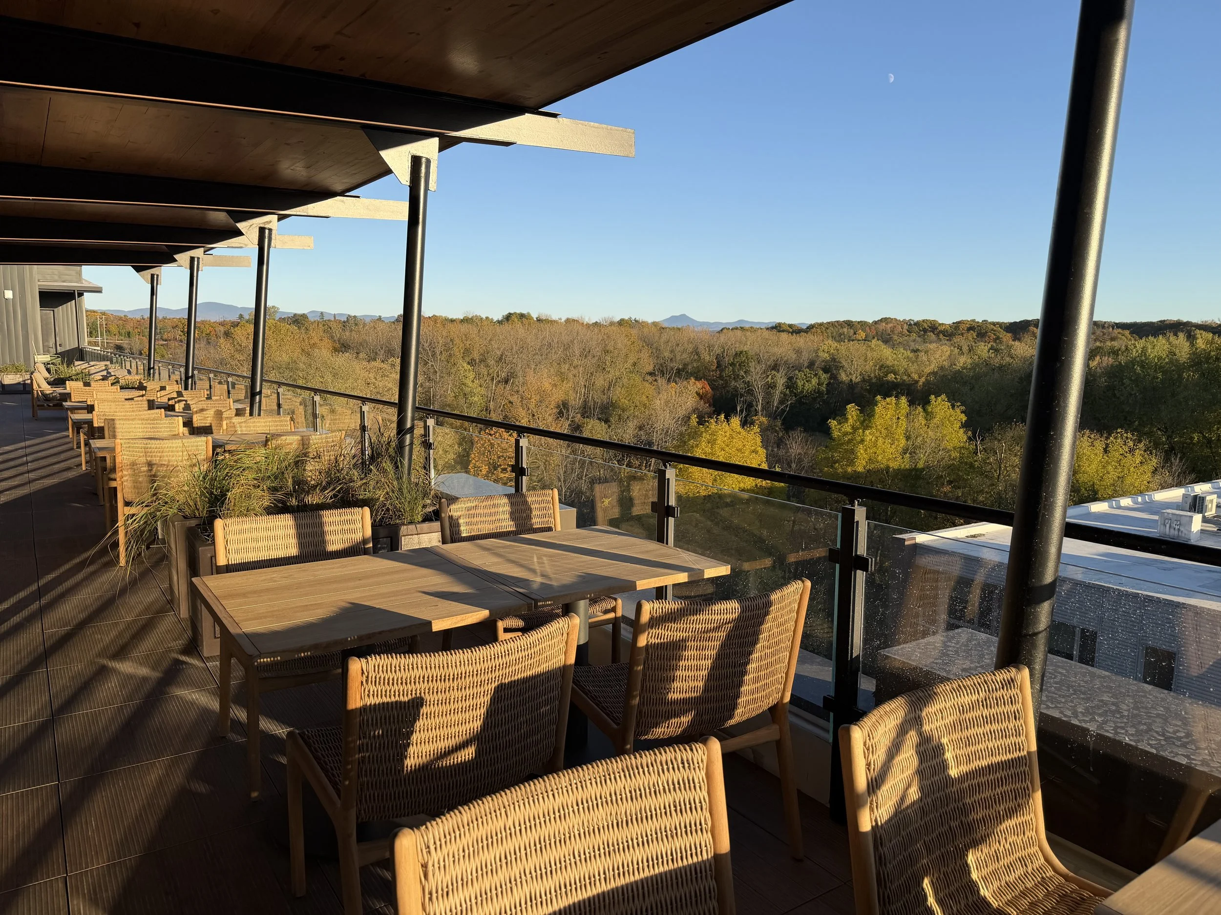 Outdoor balcony with wooden tables and wicker chairs, overlooking a forest and distant mountains under a clear blue sky, with a visible moon.