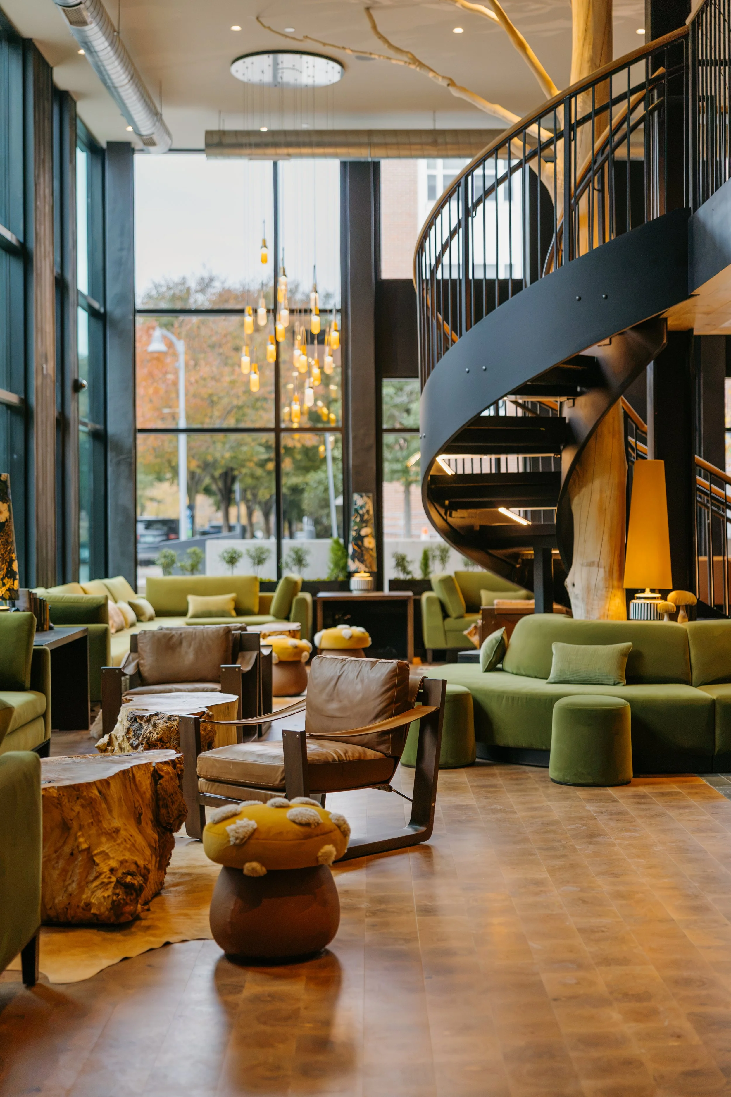 The image shows the interior of a modern, stylish lounge area with wooden accents, large windows, green sofas, armchairs, and abstract wood sculptures. There are pendant lights hanging from the ceiling and a spiral staircase with black railings.