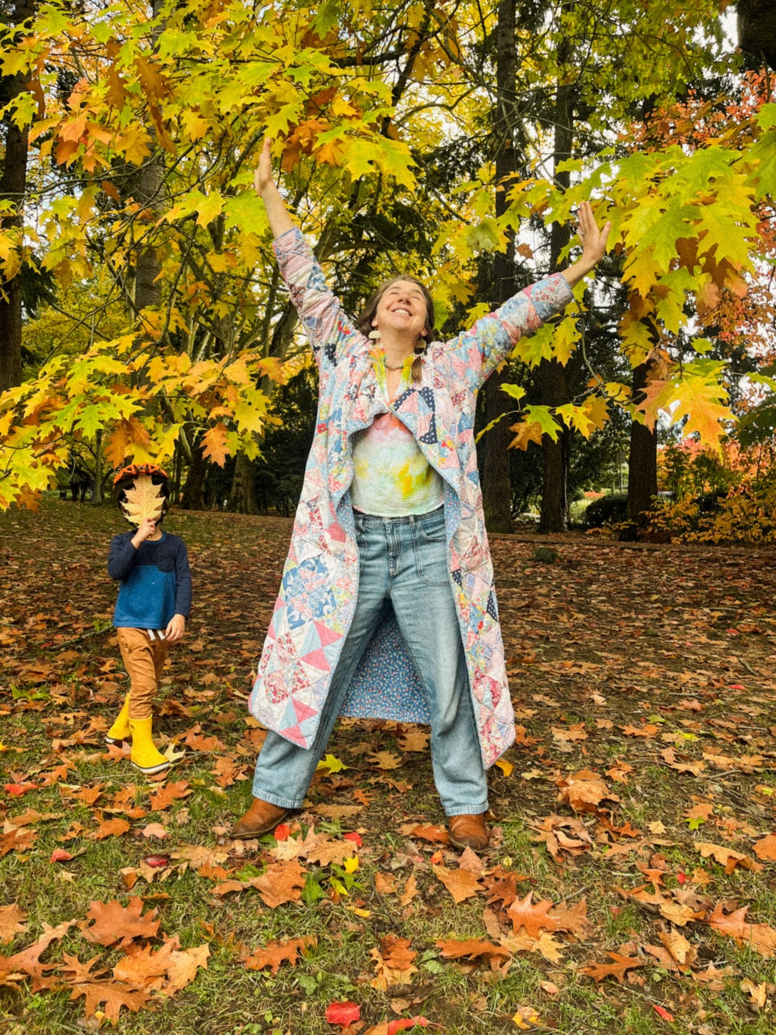 A woman standing with her arms raised in a park surrounded by colorful fall leaves, smiling happily. A child in the background wearing yellow rain boots and holding a large leaf over their face.