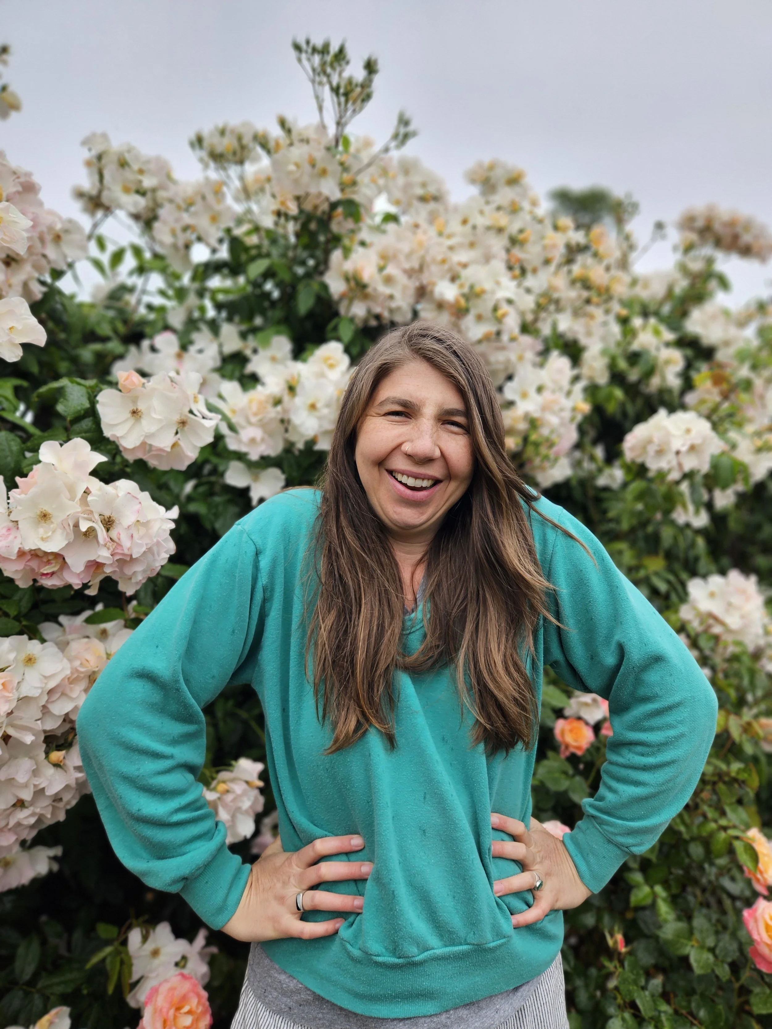 A woman with long brown hair, smiling and wearing a turquoise hoodie, standing outdoors in front of a large flowering shrub with white and pink blossoms.