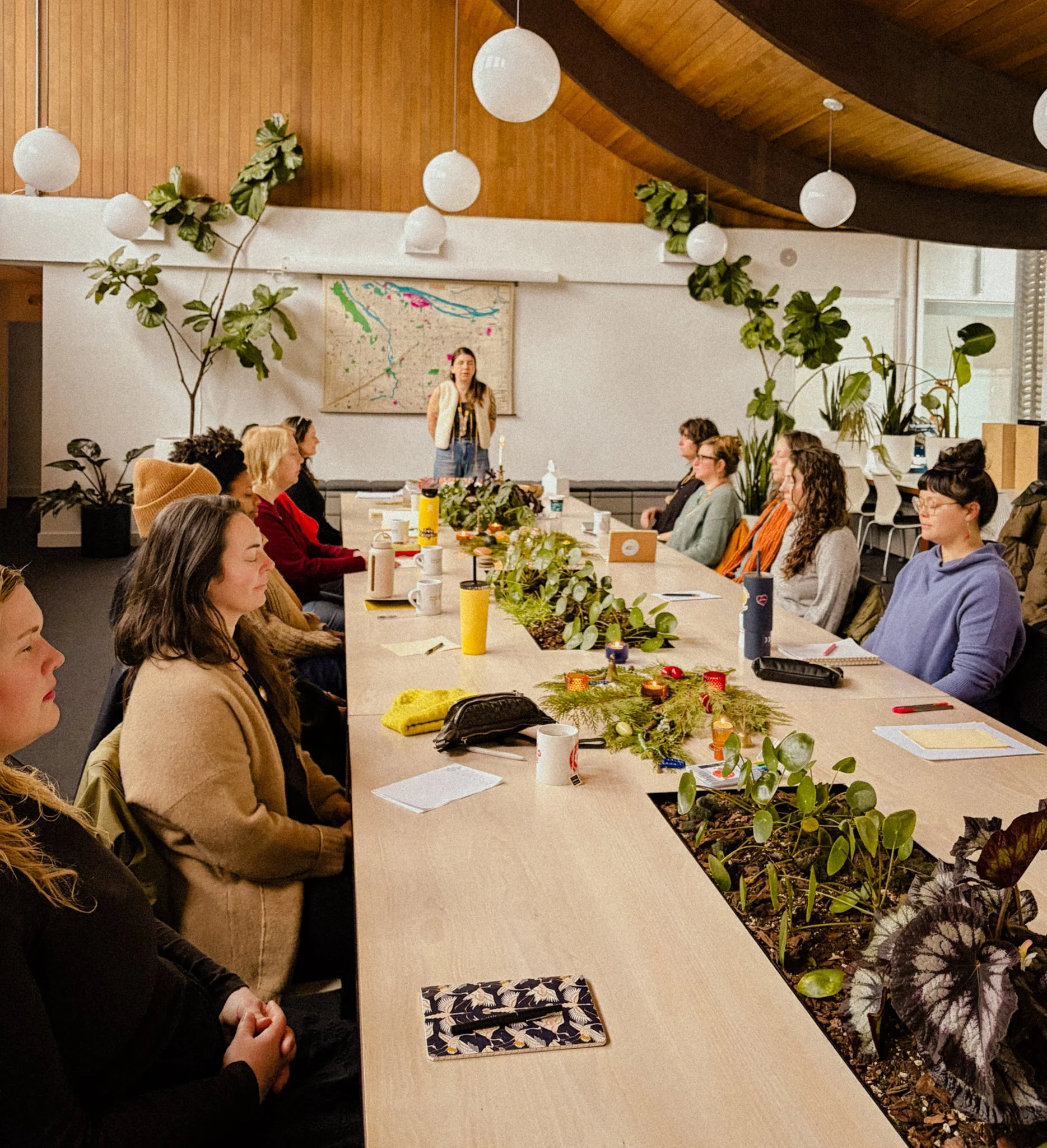 People sitting around a long table with plants as a centerpiece, in a room with wooden ceiling and hanging round lights, a person standing at the end appears to be giving a speech or presentation.