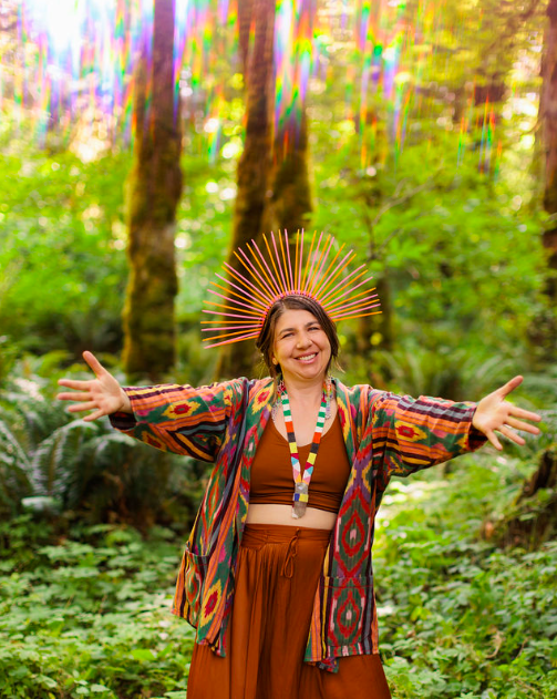 A woman standing in a lush green forest wearing a colorful patterned kimono and brown crop top, with a beaded necklace and a radiant smile, extending her arms outward. She has an elaborate headpiece made of thin, glowing lines radiating outward, resembling a crown or halo.